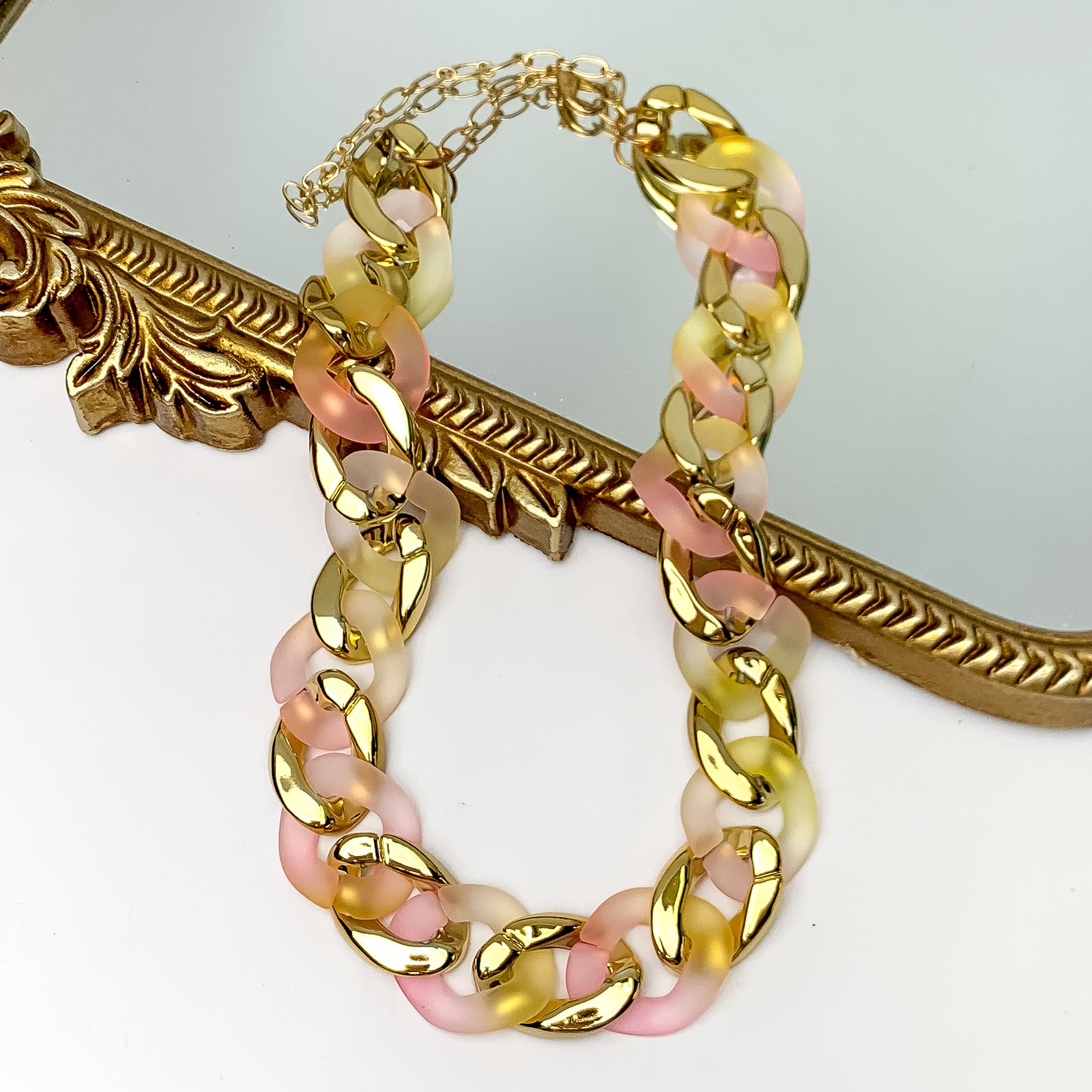 Pictured partially on a gold mirror on a white background is a chain link necklace in gold and pink. 