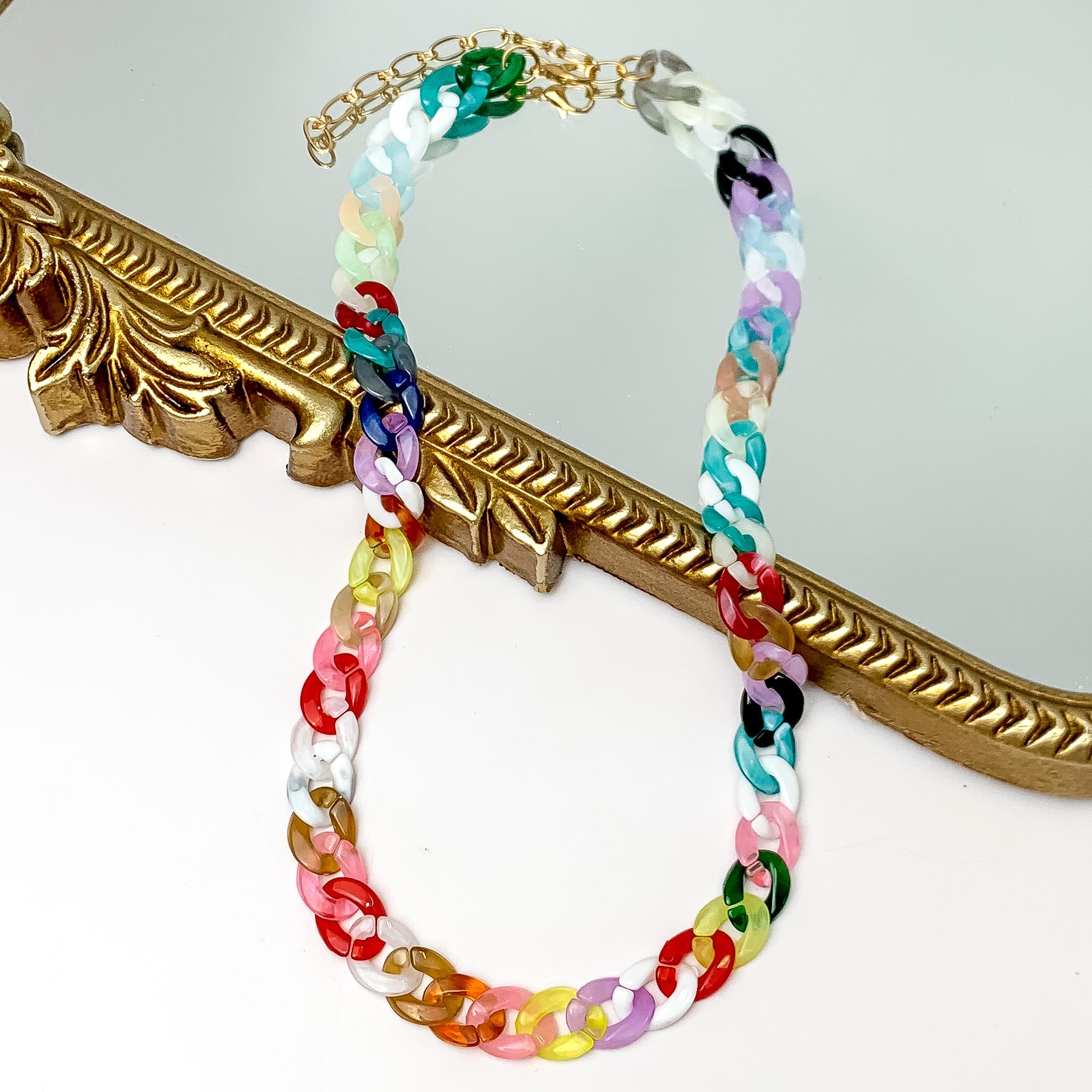 Pictured partially on a gold mirror on a white background is a chain link necklace in multicolor. This necklace includes pinks, blues, reds, and white. 
