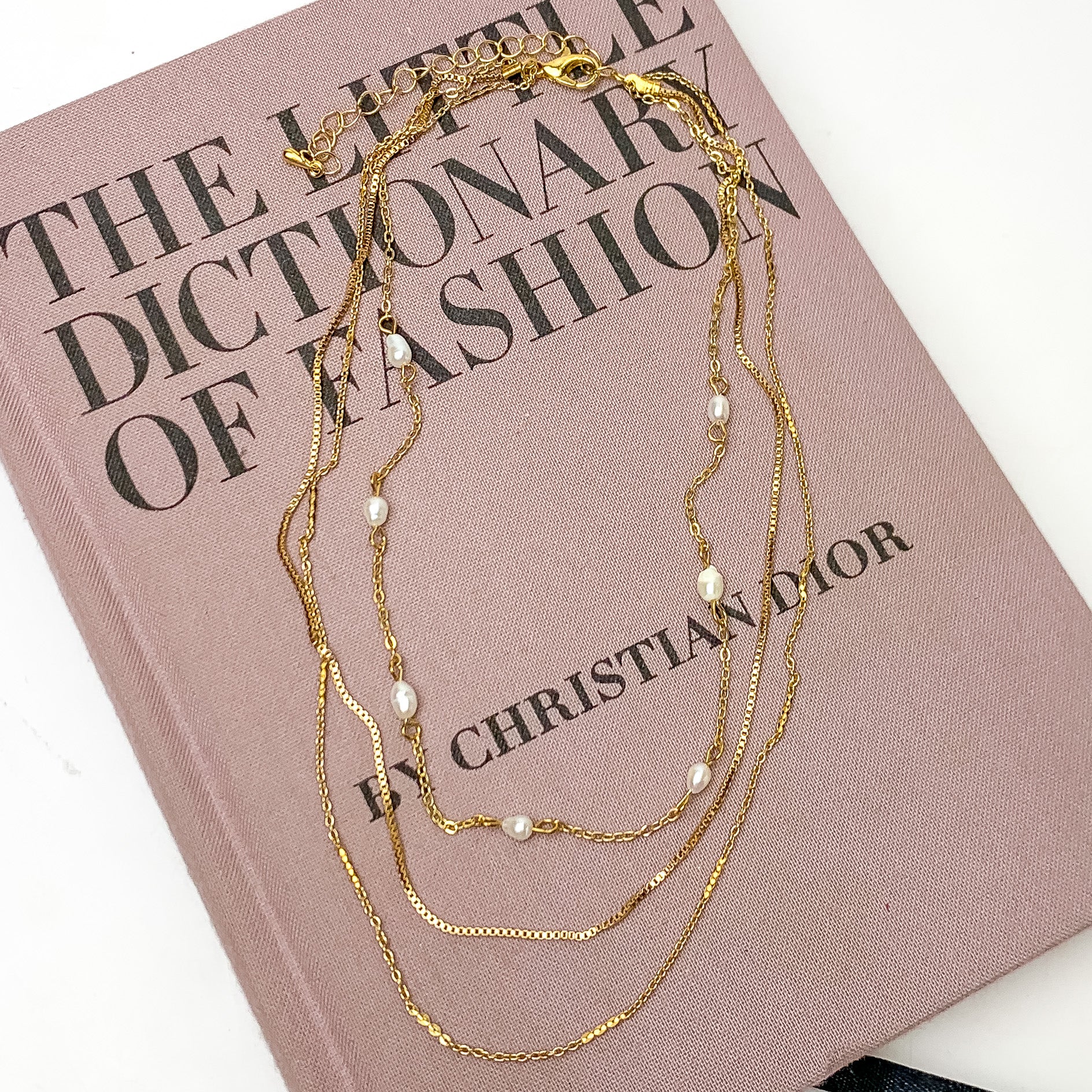 Three Strand Gold Tone Necklace With Pearl Accents. Pictured on a white background with the necklace laying on a pink book.