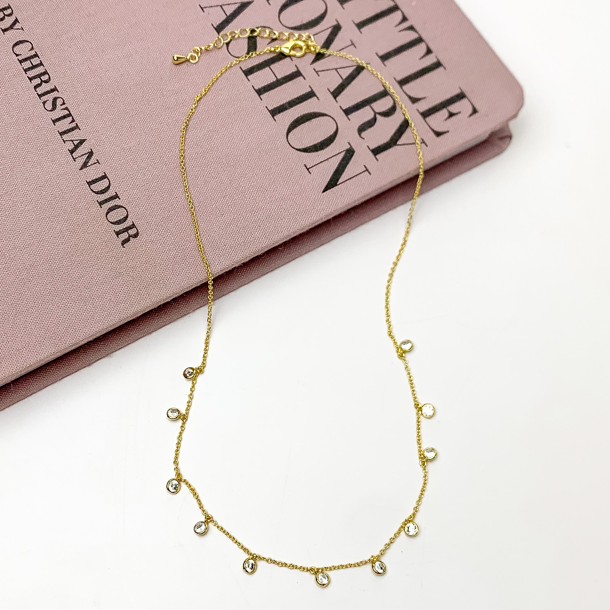 Gold Tone Simple Necklace With Multiple Clear Crystals. Pictured on a white background with the top of the necklace laying on a pink book.