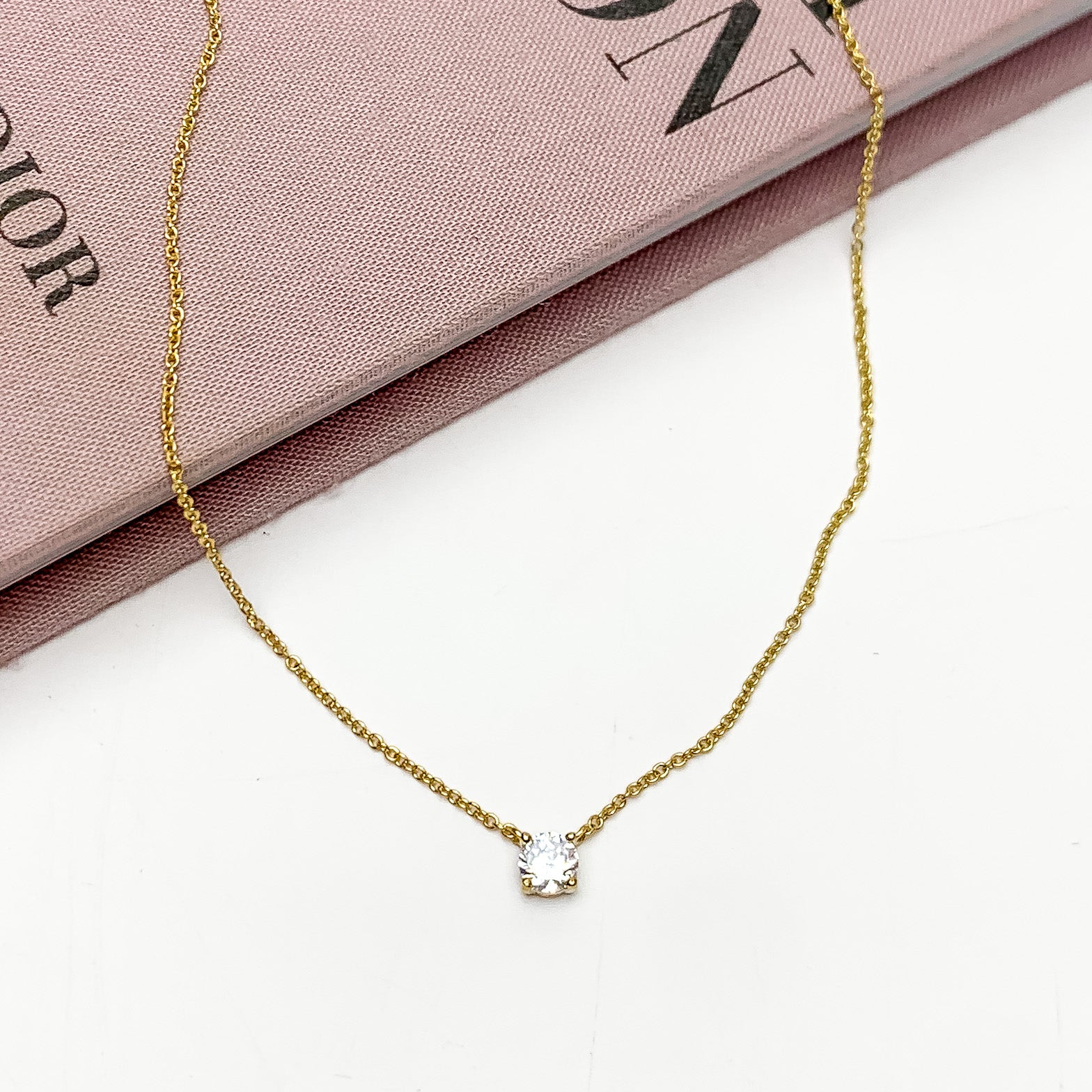 Gold Tone Simple Necklace With Clear Crystal. Pictured on a white background with the top of the necklace on a pink book.