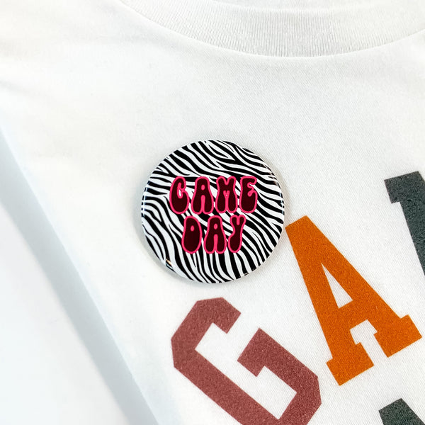Black and white zebra print button pin pictured on a white tee shirt. This pin includes the words "GAME DAY" in maroon with a pink outline.  