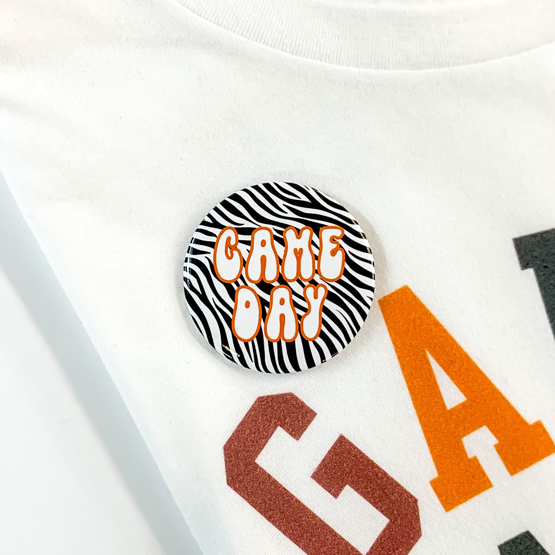 Black and white zebra print button pin pictured on a white tee shirt. This pin includes the words "GAME DAY" in white with an orange outline.  