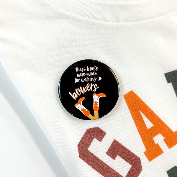 Black button pin pictured on a white tee shirt. This pin includes the words "these boots were made for walking to bowers" in white with orange boots at the bottom.  