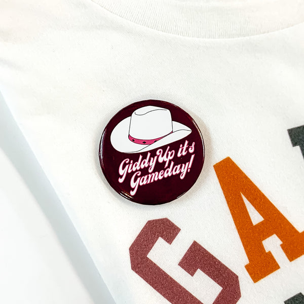 Dark maroon button pin pictured on a white tee shirt. This pin includes a white cowboy hat and the words "Giddy Up It's Gameday!" in white with a pink outline. 