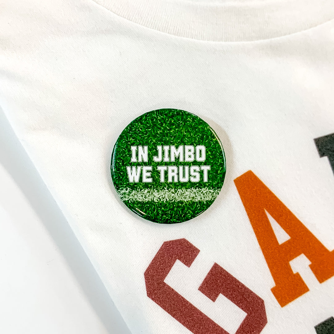 Green button pin pictured on a white tee shirt with a disco ball print. This pin says "IN JIMBO WE TRUST" in white.  