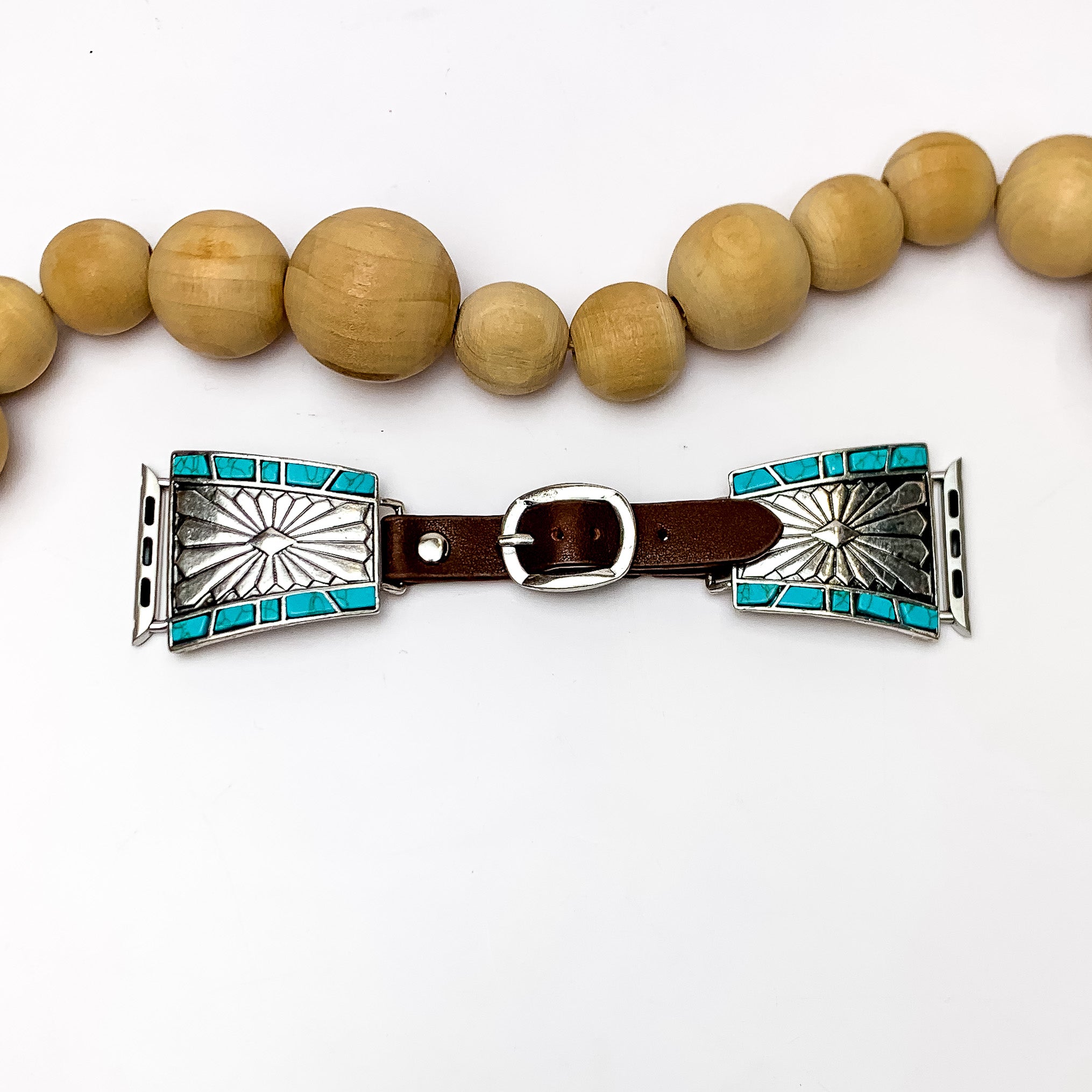 Brown Watch Band with Silver Tone Designs and Turquoise Stones. Pictured on a white background with wood beads above the band for decoration. 
