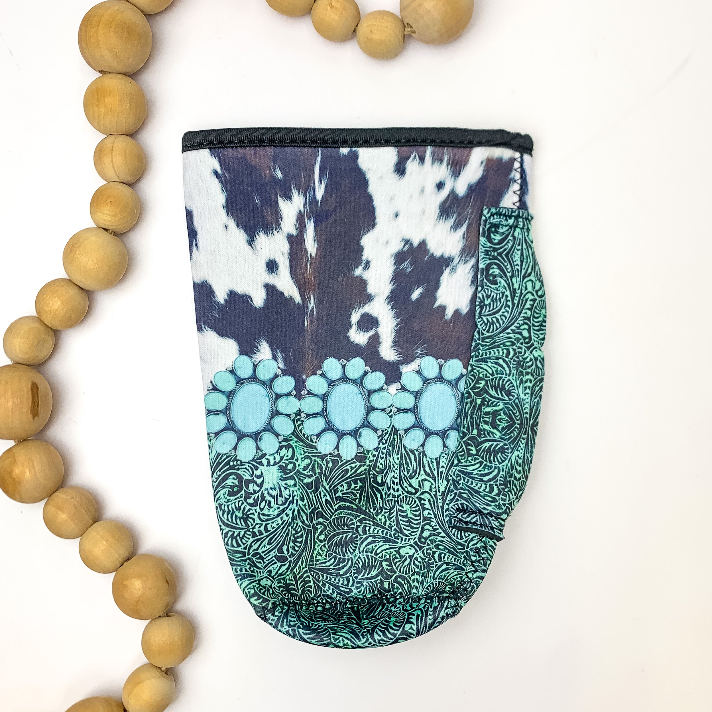 Cow Print Tumbler Drink Sleeve With Turquoise Designs. Pictured on a white background with wood beads to the left of the koozie for design.