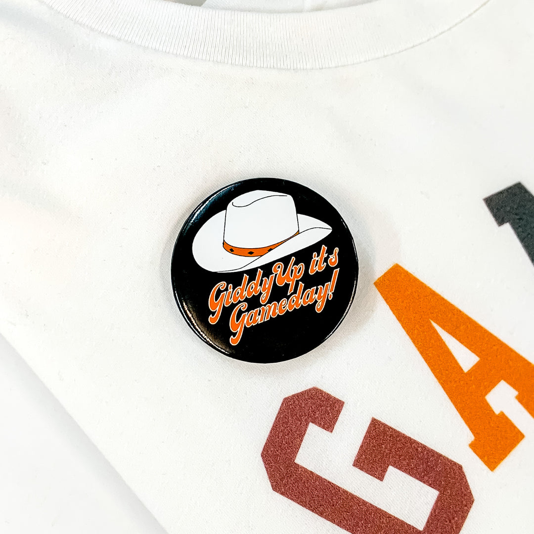 Black button pin pictured on a white tee shirt. This pin includes a white cowboy hat and the words "Giddy Up It's Gameday!" in orange. 