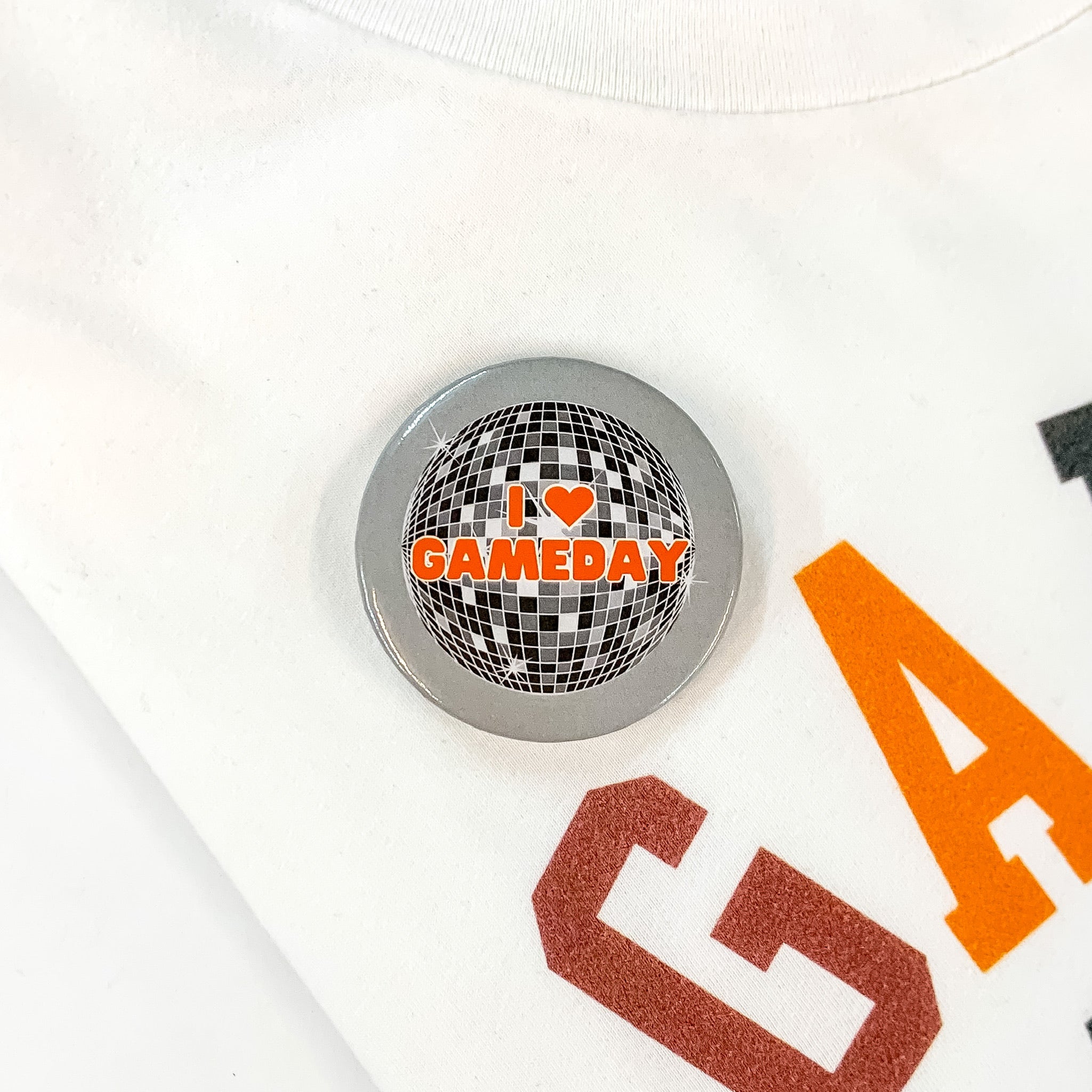 Grey button pin pictured on a white tee shirt with a disco ball print. This pin says "I love GAMEDAY" with a heart for the word love in orange.  