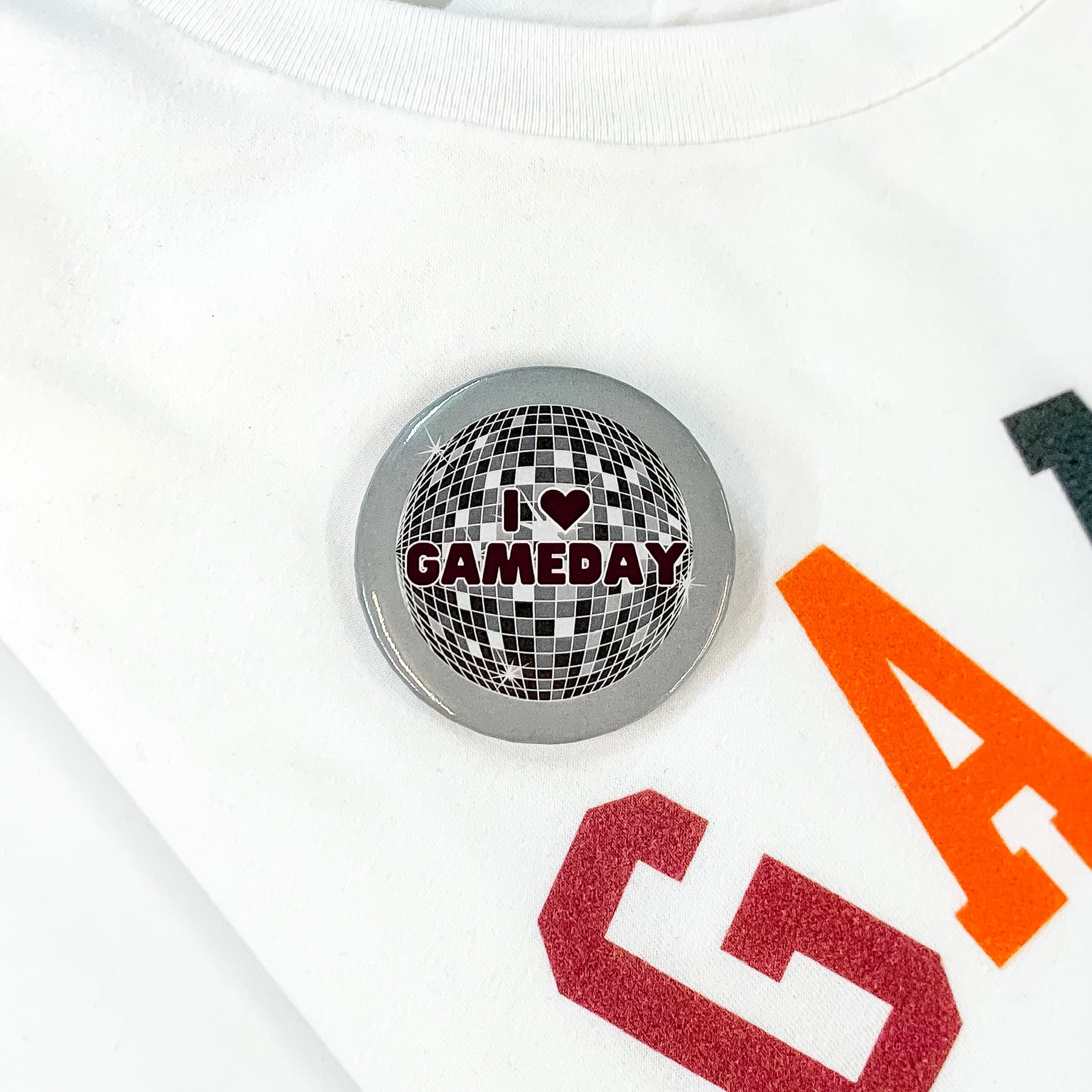 Grey button pin pictured on a white tee shirt with a disco ball print. This pin says "I love GAMEDAY" with a heart for the word love in maroon.  