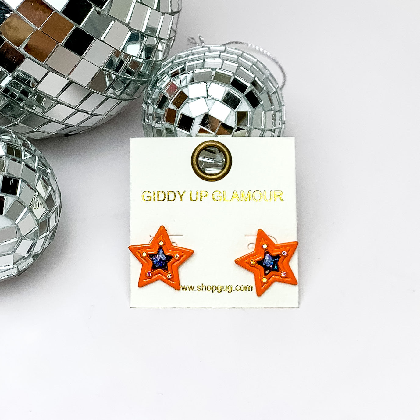 Orange, star shaped earrings with a blue center on a white earring holder. These earrings are pictured on a white background with disco balls in the top left corner. 