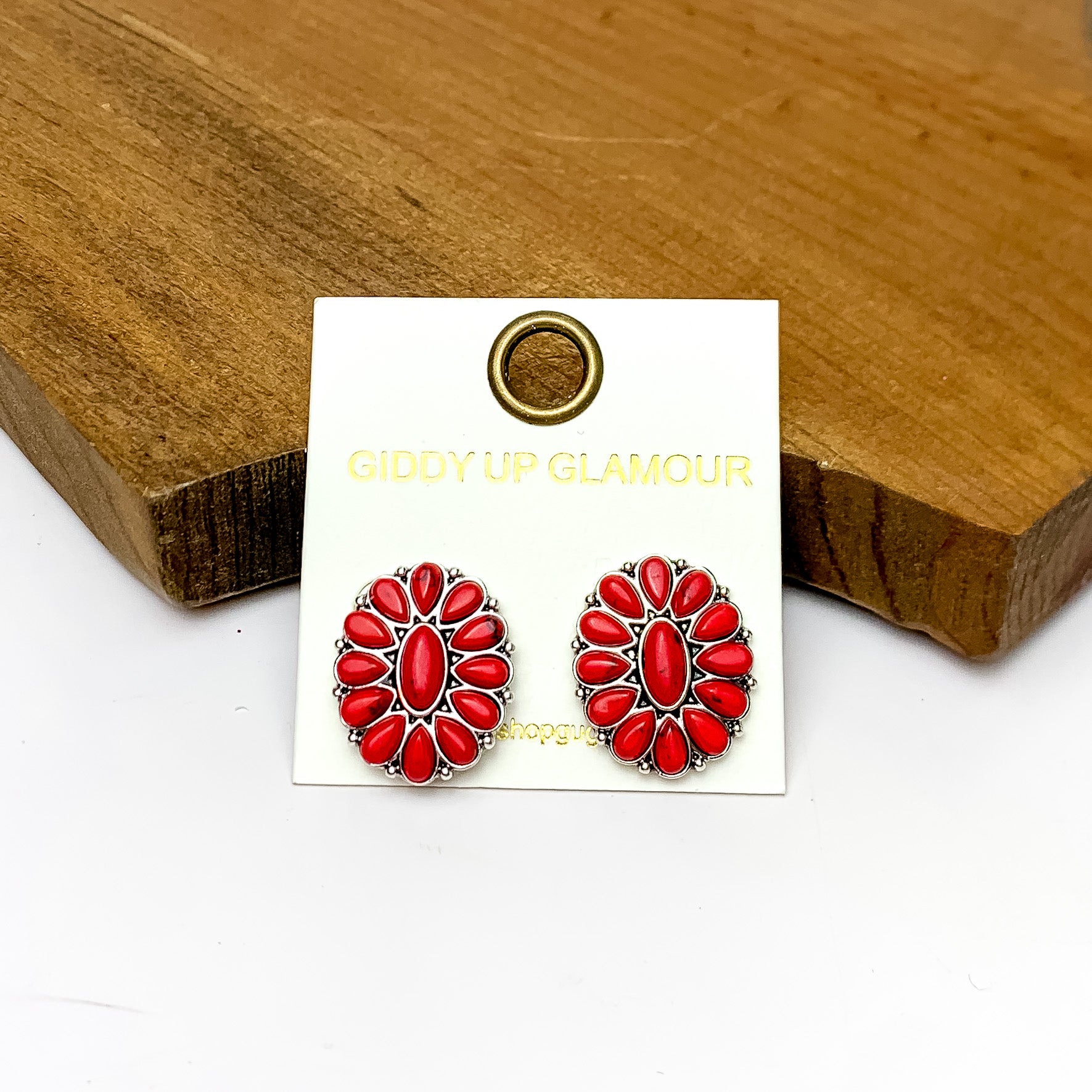 Silver Tone Concho Cluster Oval Earrings in Red. Pictured on a white background with the earrings laying against a wood piece.