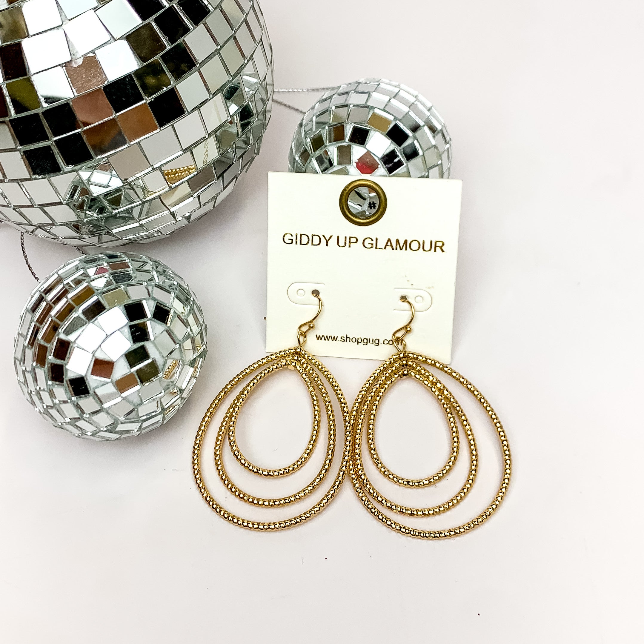 Really Radiant Triple Open Teardrop Earrings in Gold Tone. Pictured on a white background with disco balls in the top left corner.