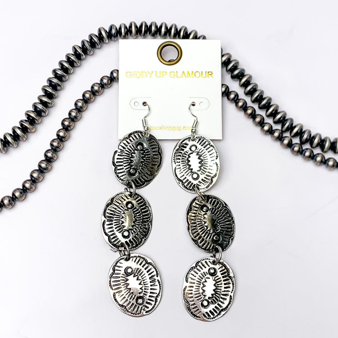 Western Oval Design Dangle Earrings in Silver Tone. Pictured on a white background with strands of silver beads behind the earrings for decoration.
