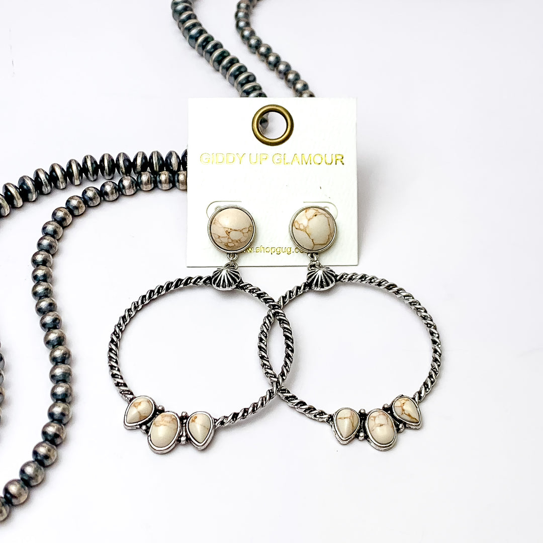 Circle, ivory stone post earrings with a silver circle drop pendant. At the bottom of the circle there are three different shaped stones in ivory. These earrings are pictured on a white background with silver beads on the left side bottom of the picture. 