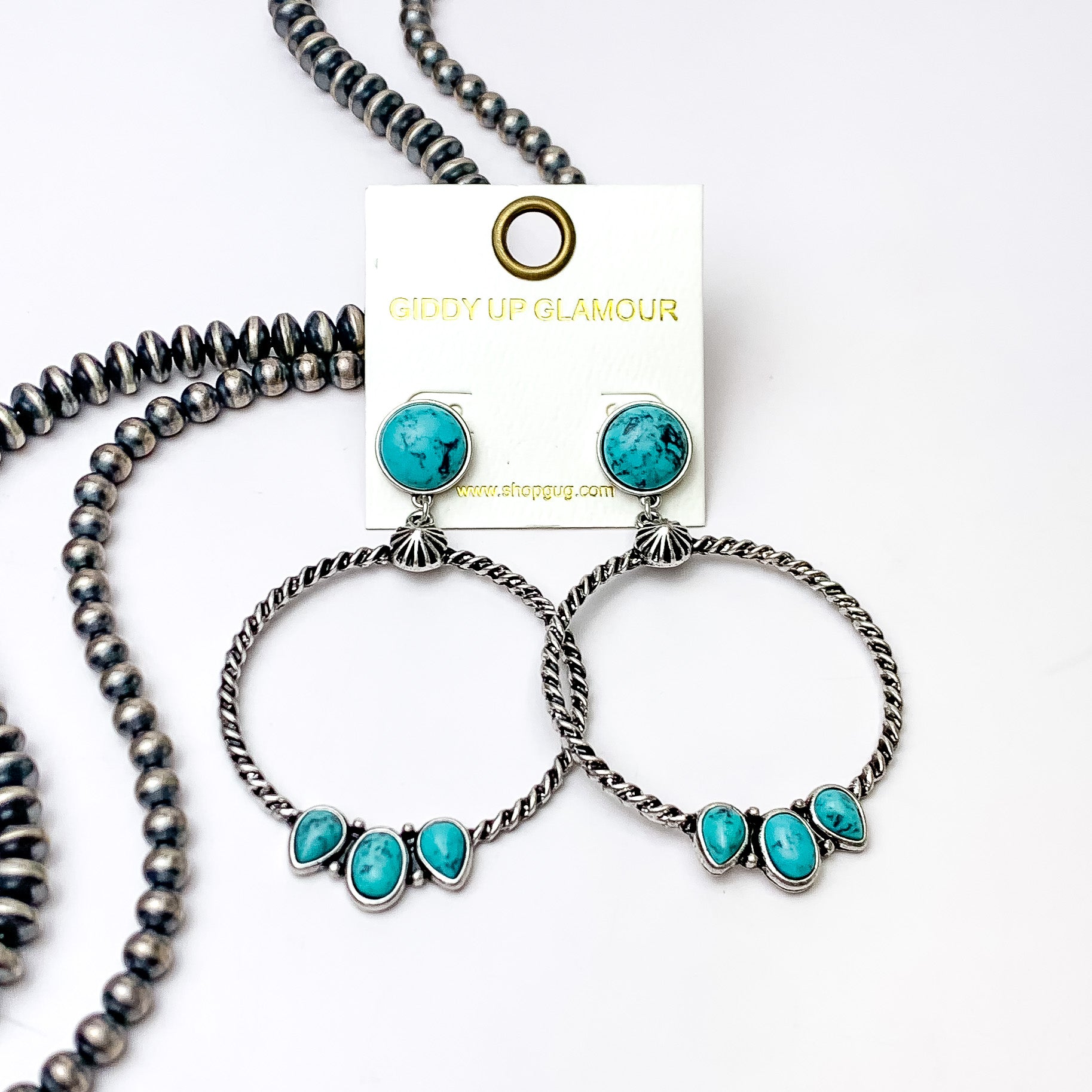 Circle, turquoise stone post earrings with a silver circle drop pendant. At the bottom of the circle there are three different shaped stones in turquoise. These earrings are pictured on a white background with silver beads on the left side bottom of the picture. 