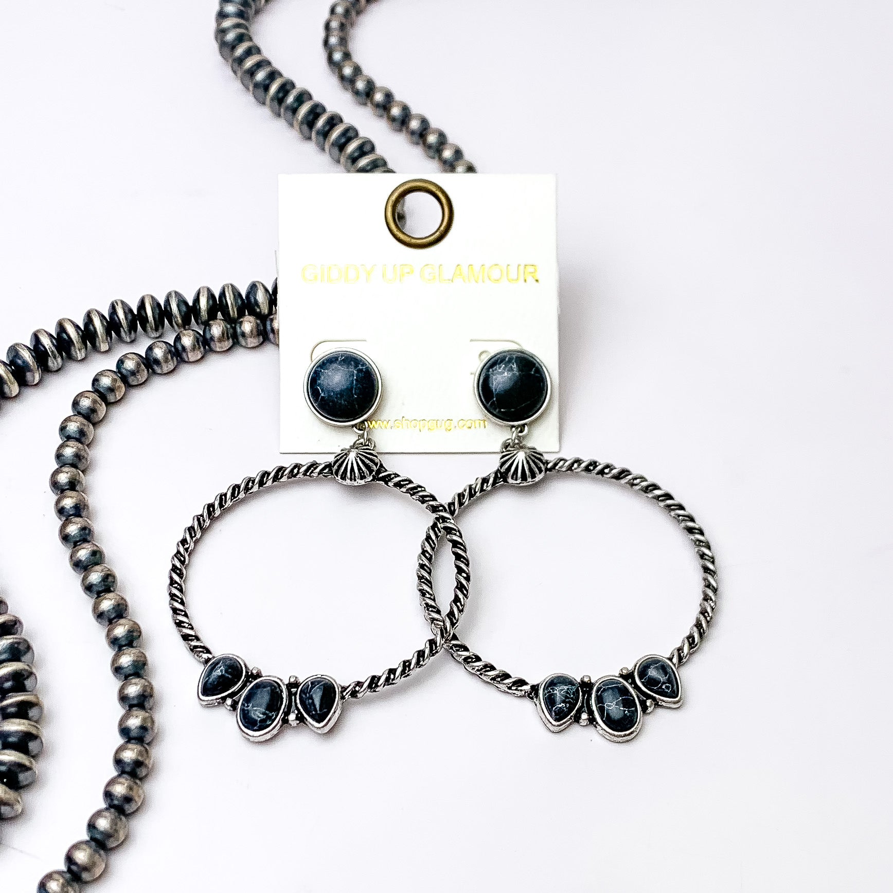Circle, black stone post earrings with a silver circle drop pendant. At the bottom of the circle there are three different shaped stones in black. These earrings are pictured on a white background with silver beads on the left side bottom of the picture. 
