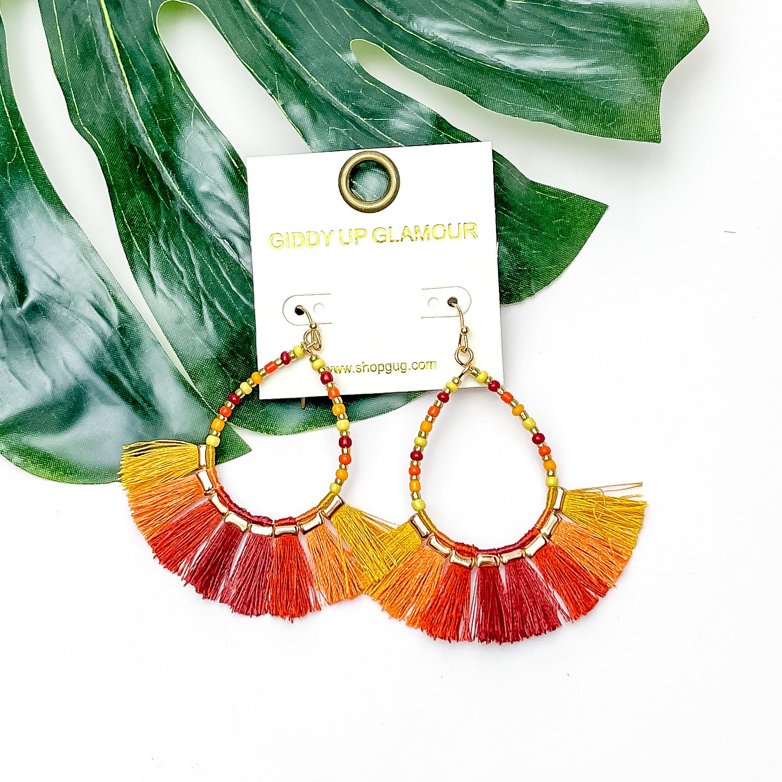 Beaded Open Teardrop Earrings With Fringe Bottom in Orange Tones. Pictured on a white background with a leaf behind the earrings.