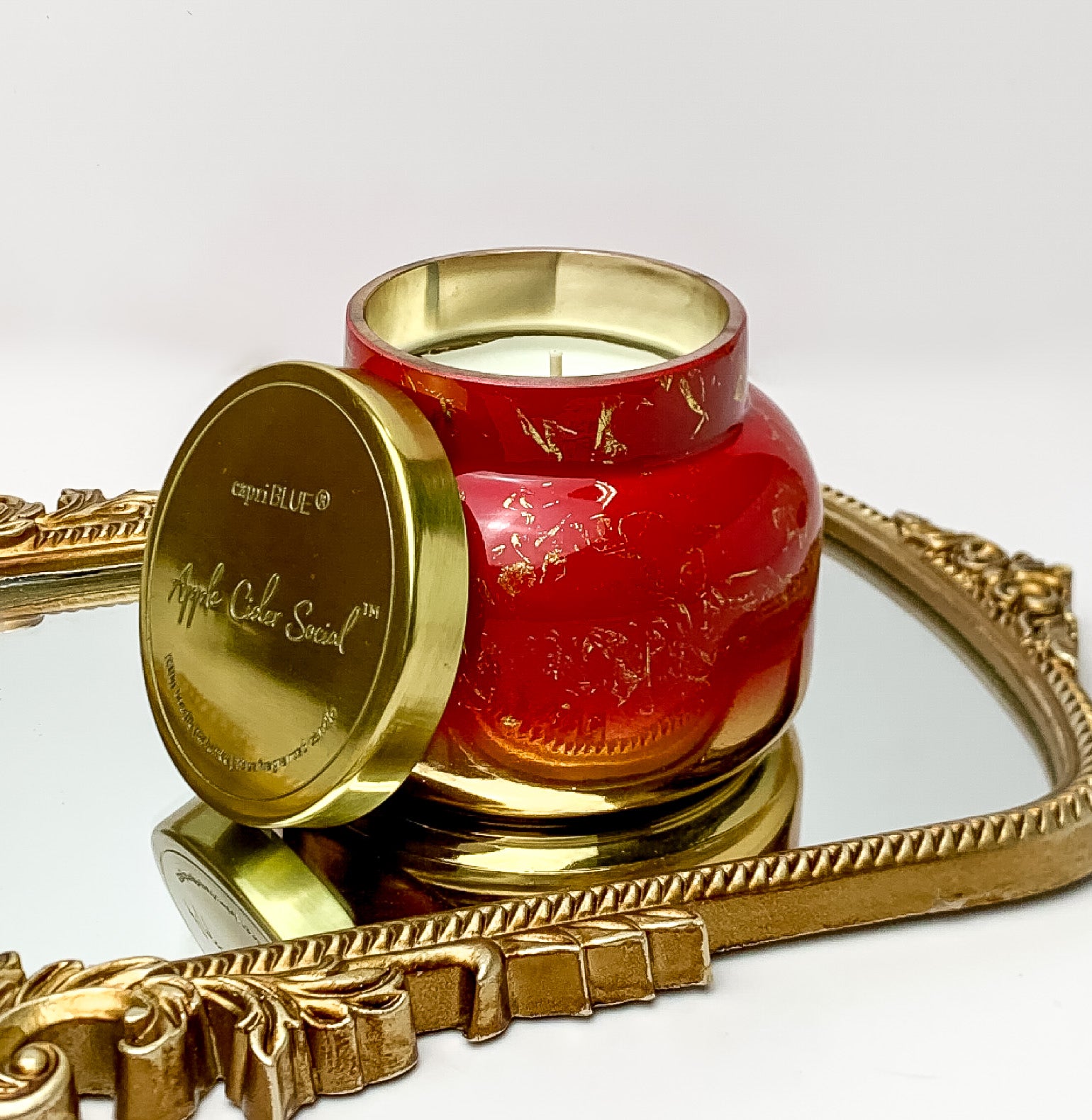 Capri Blue | 19 oz. Jar Candle in Glimmer Signature Jar Candle | Apple Cider Social. This candle is red and gold with a gold lid. The candle is sitting on a gold framed mirror with a white background. 