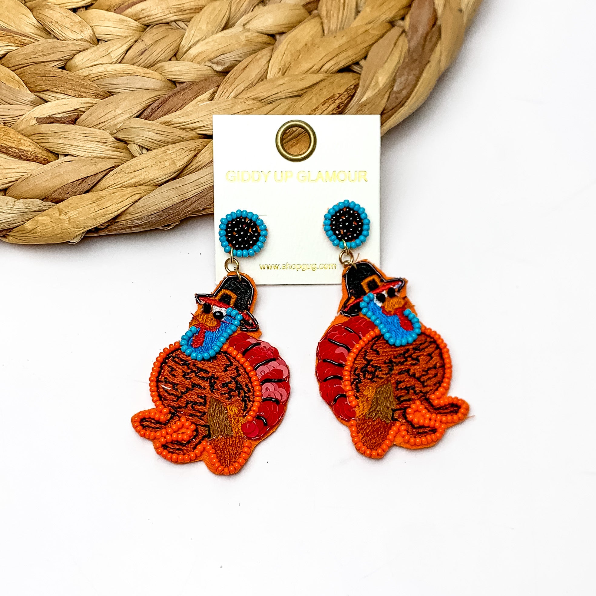 Fall Time Beaded Turkey Earrings in Brown, Red, and Orange. Pictured on a white background with the earrings laying against a wicker piece.
