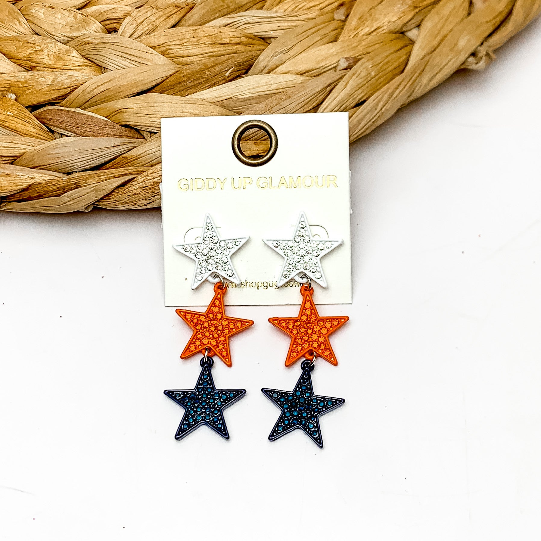 Star Shaped Three Tier Earrings in White, Orange, and Navy. The earrings are laying against a woven piece. The background of the picture is white.