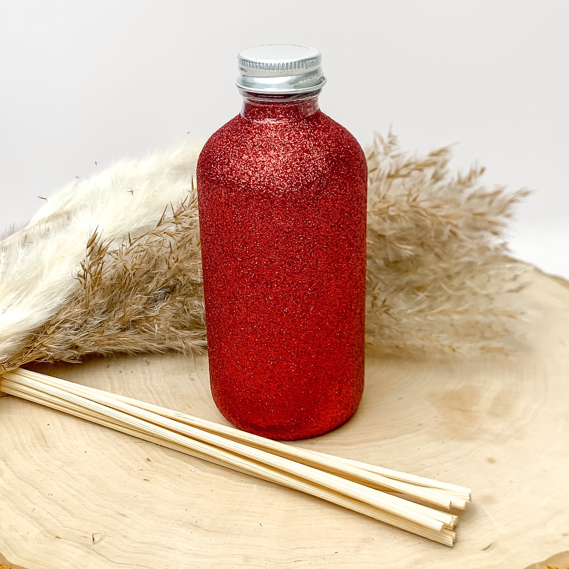Capri blue red sparkly reed diffuser. There are wood sticks in front of the diffuser to put in the bottle. The bottle is sitting on a wood piece with feathers behind it. The background of the picture is white. 