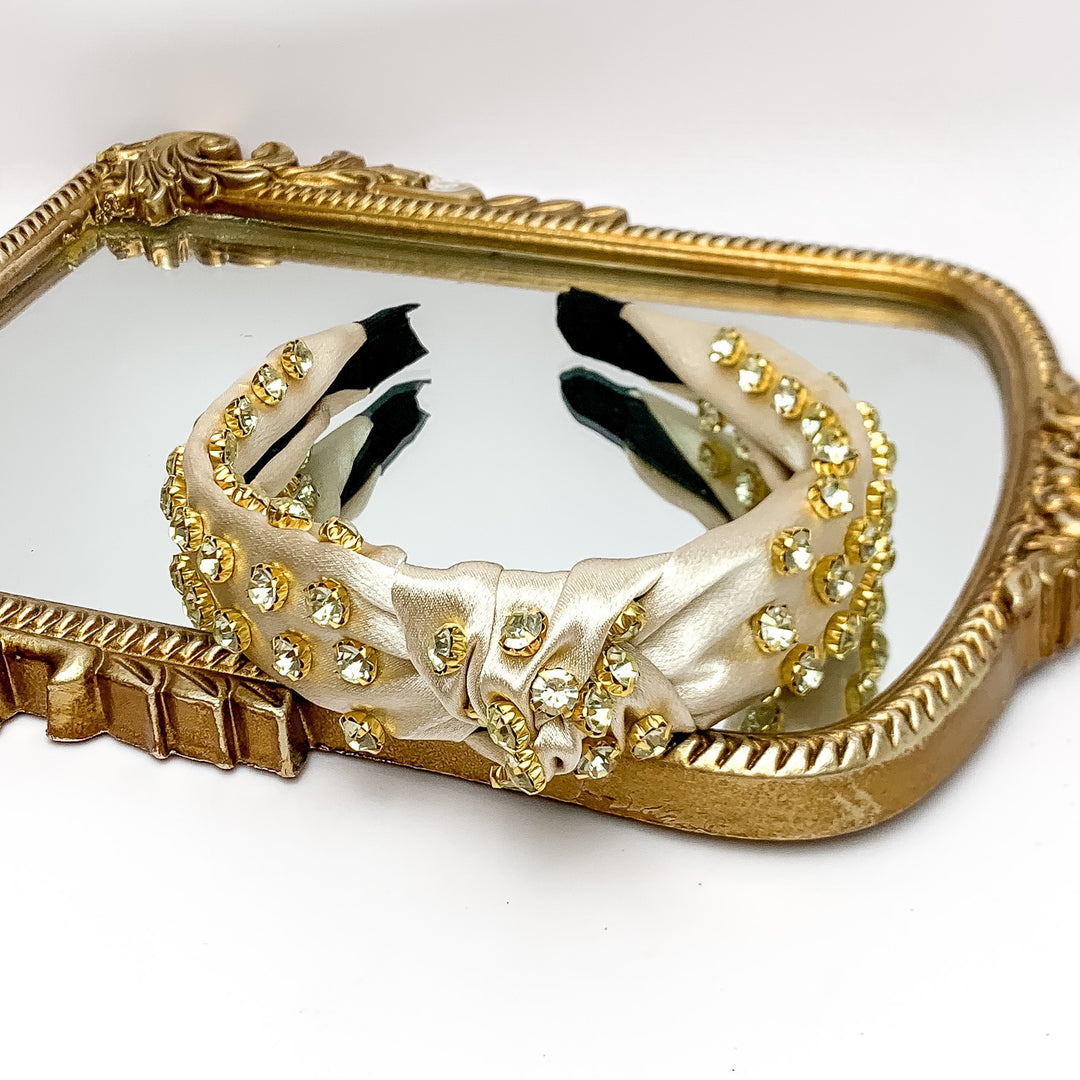 Crystal Detailed Satin Knot Headband in Champagne. Pictured on a white background with the headband on a gold trimmed mirror.