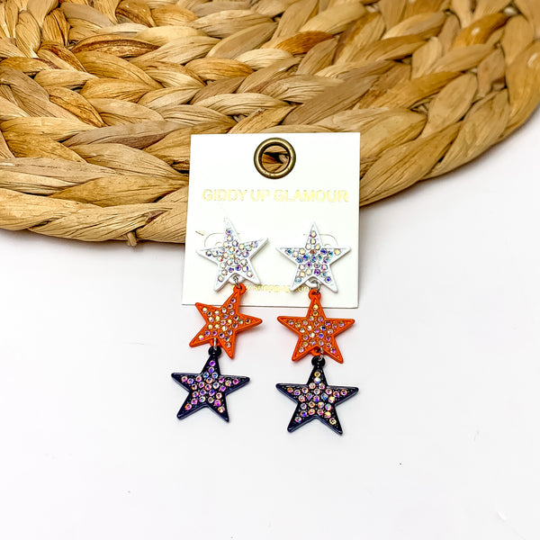 Star Shaped Three Tier Earrings With Clear Crystals in White, Orange, and Navy. Pictured on a white background with the earrings laying against a wicker piece.