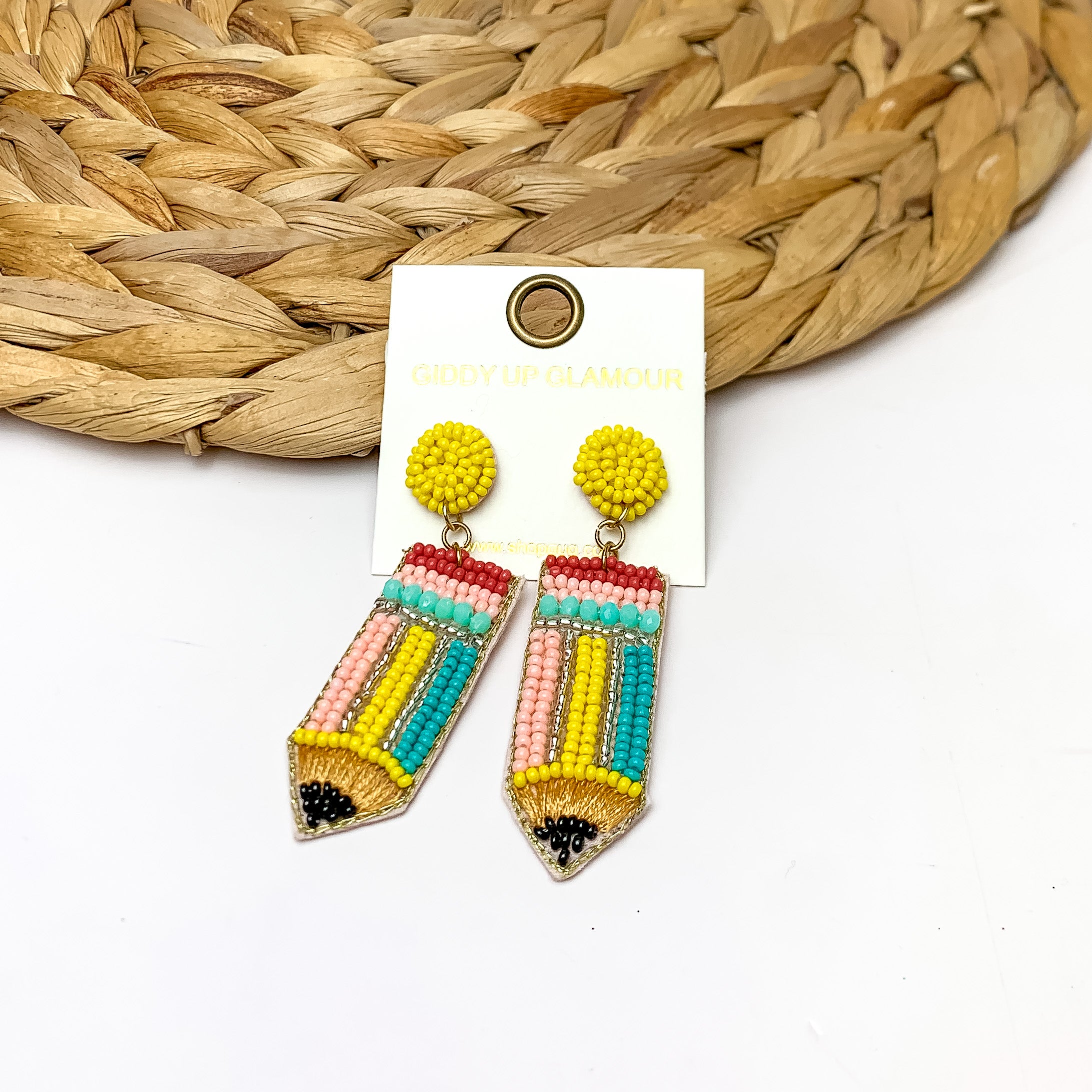 Beaded Pencil Earrings in Multicolor. Pictured on a white background with the earrings laying against a wicker piece.