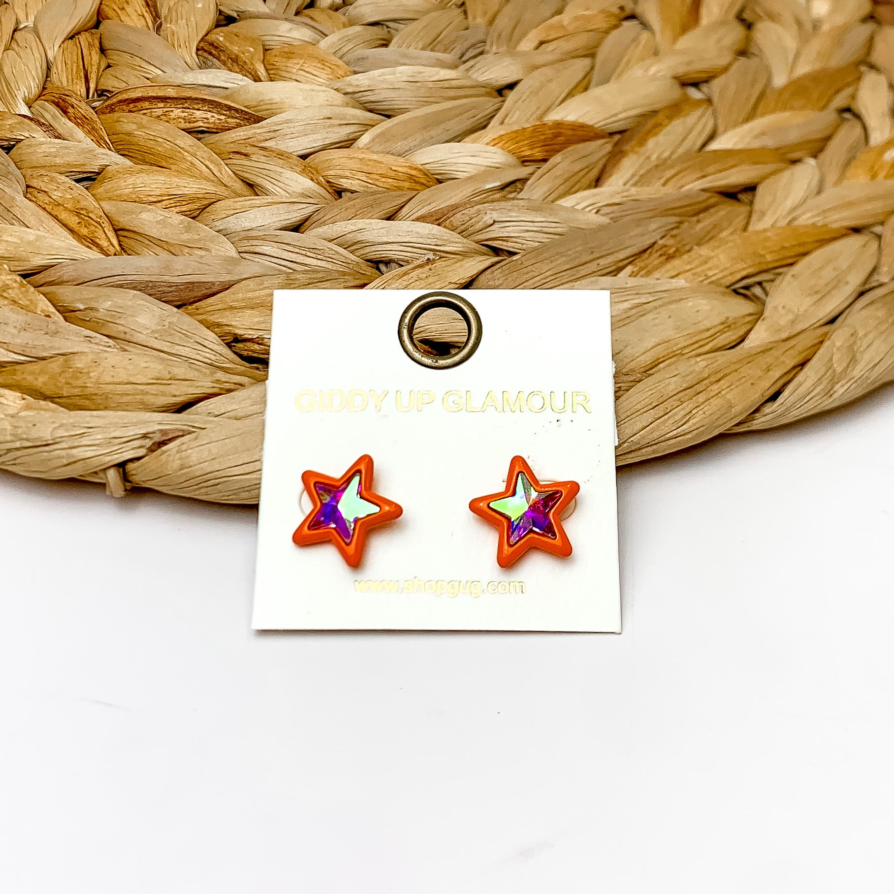 Star Stud Earrings in Orange With AB Crystals. Pictured on a white background with the earrings laying against a wicker piece.