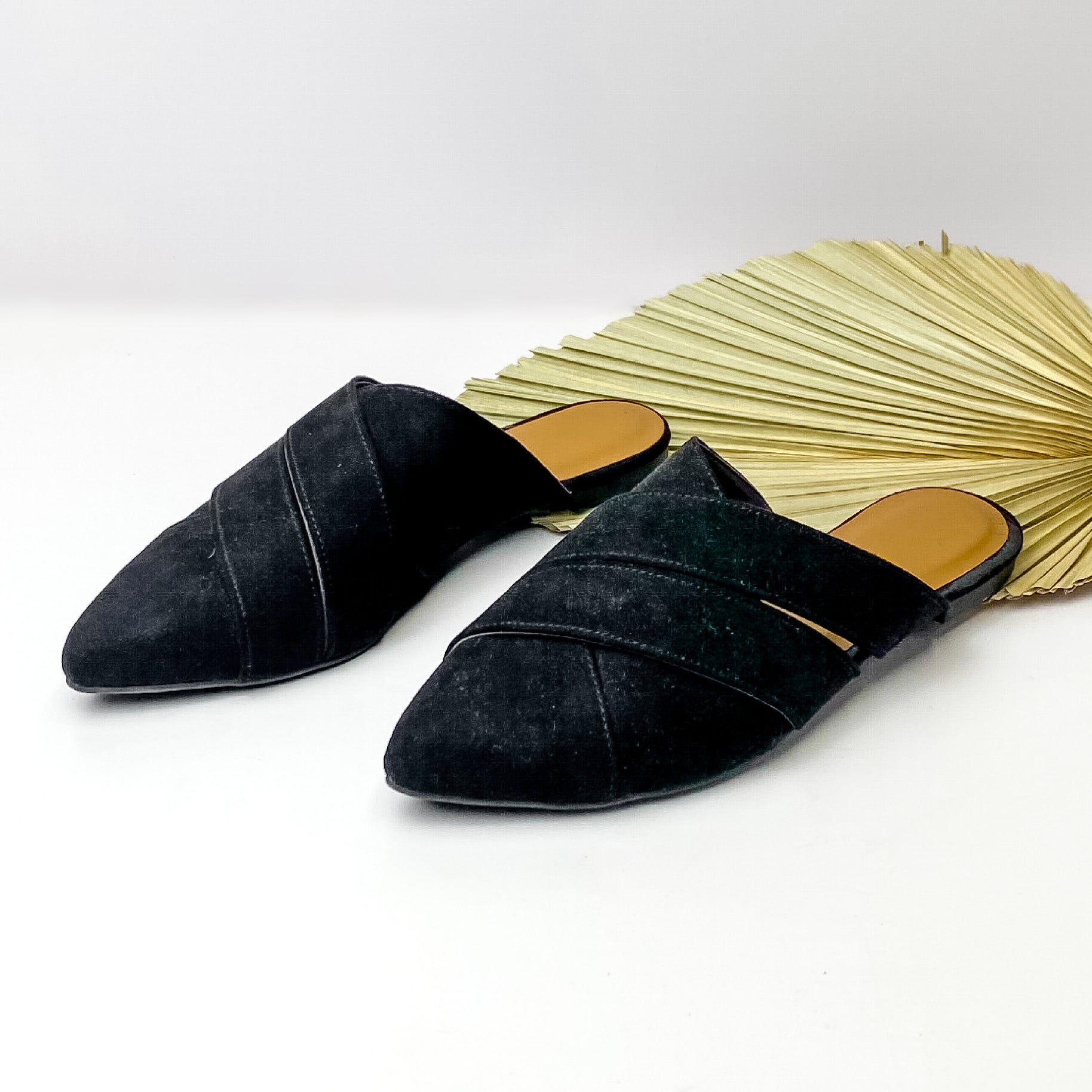 Black, pointed toe mules pictured on a white background with a green leaf on the right hand side. 