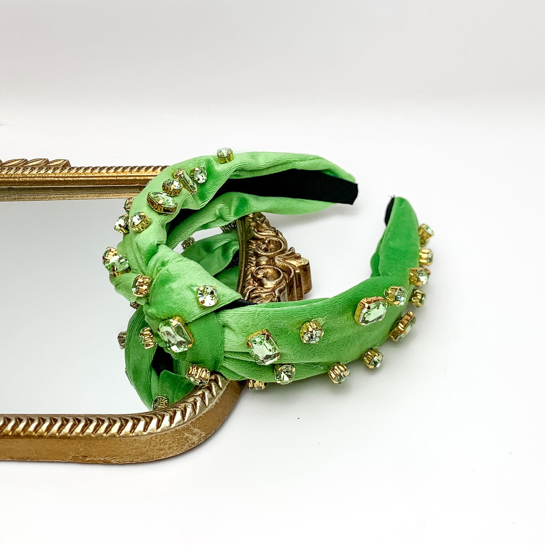 Crystal Detailed Velvet Knot Headband in Green. This headband is pictured on a white background with part of the headband on a gold trimmed mirror.