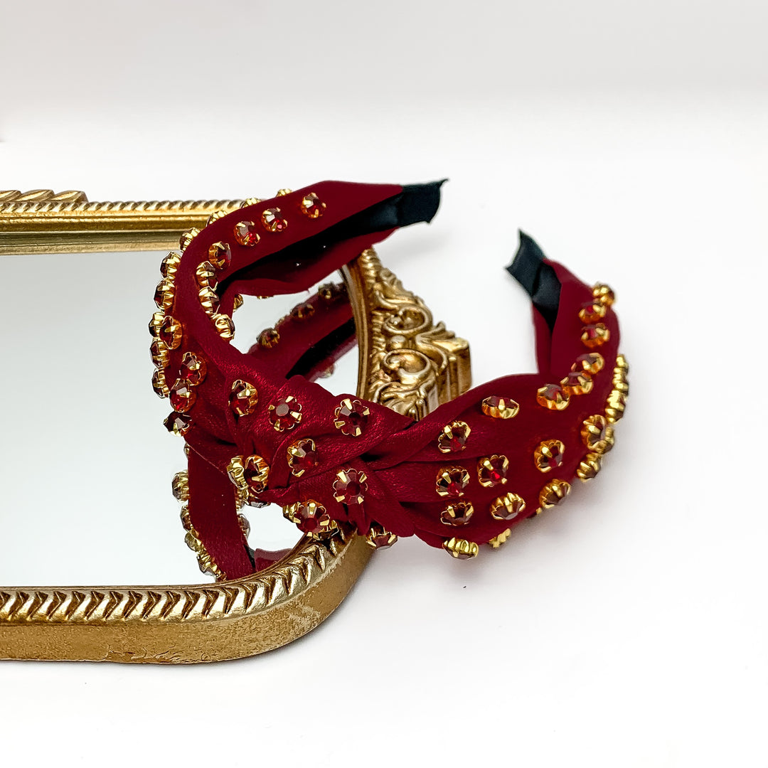 Crystal Detailed Satin Knot Headband in Maroon. Pictured on a white background with part of the headband on a gold framed mirror.