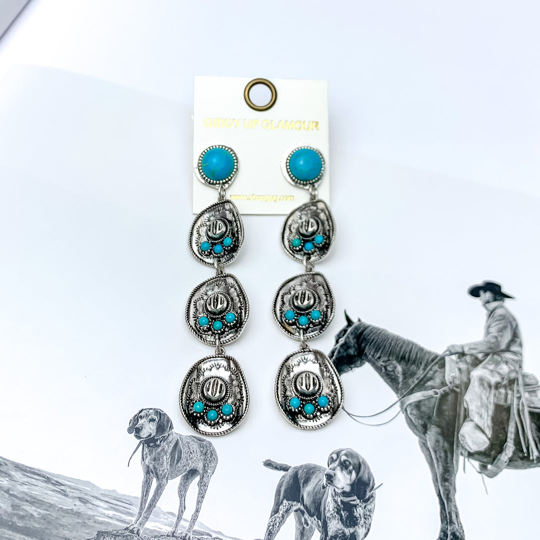 Three Tier Cowboy Hat Earrings in Silver Tone and Turquoise Blue. These cowboy hat earrings are pictured on western background. 