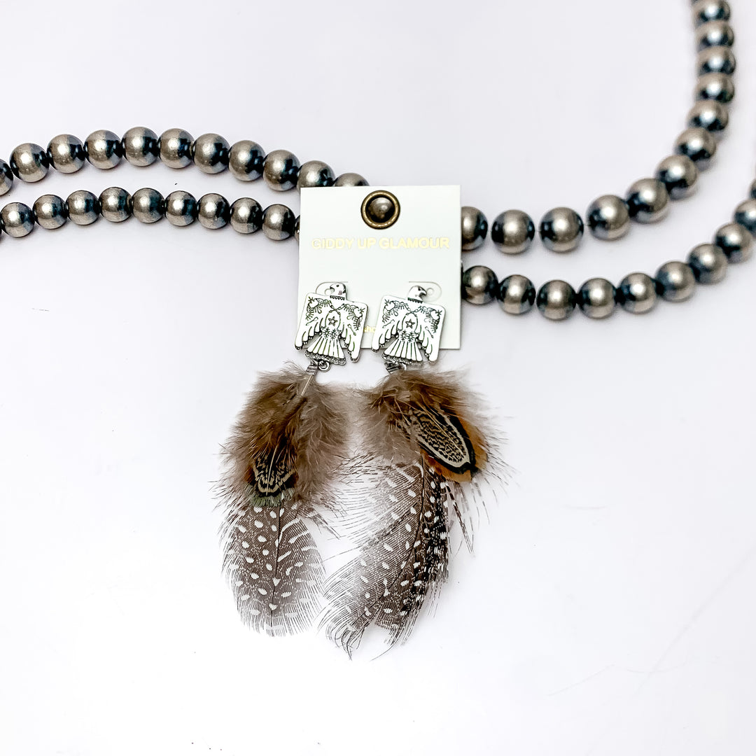 Thunder Bird Post Feather Earrings in Brown and Black. These Earrings are pictured laying against Navajo pearls with a white background.