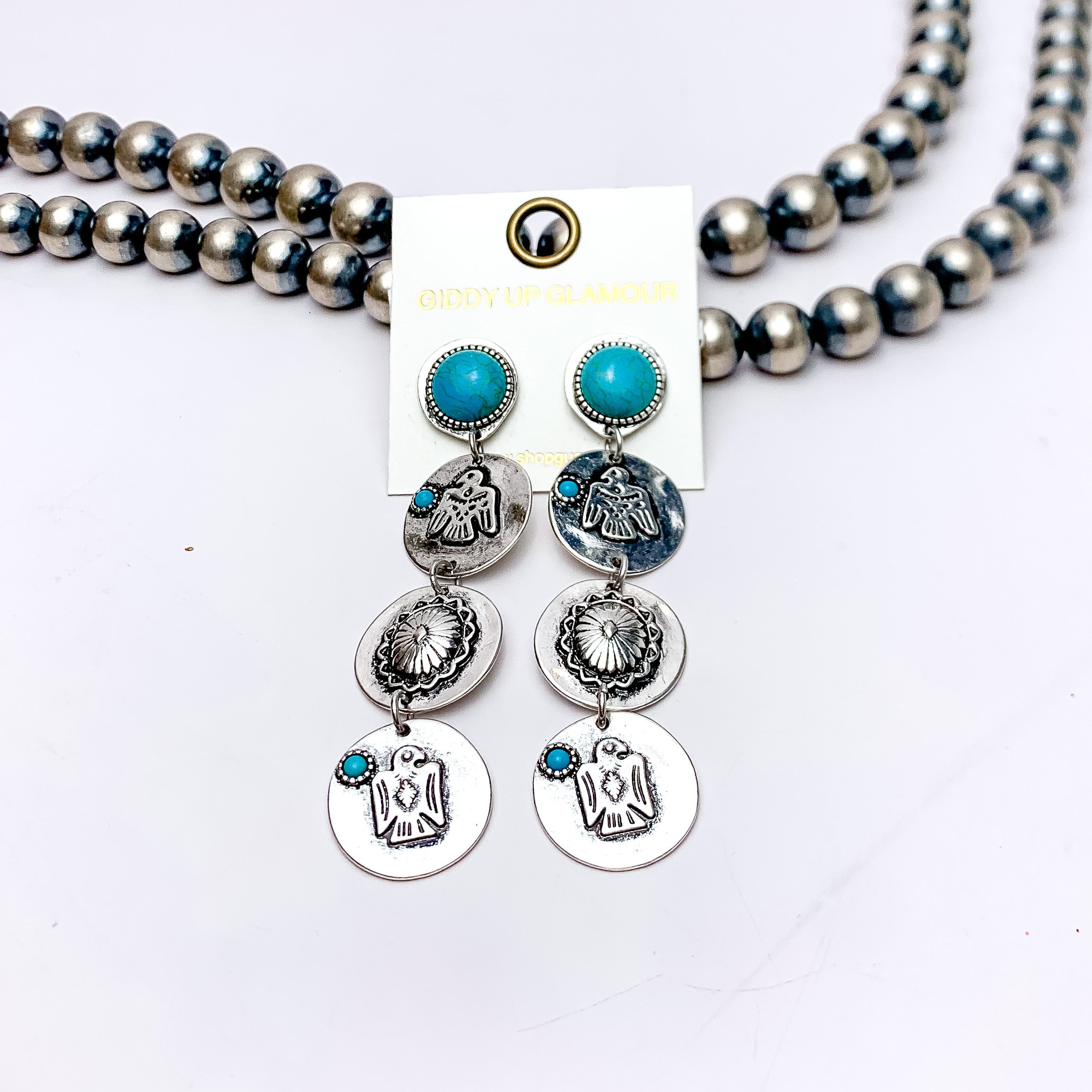 Three Tier Thunder Bird Earrings in Silver Tone and Turquoise Blue. These earrings are pictured laying against Navajo pearls and a white background.