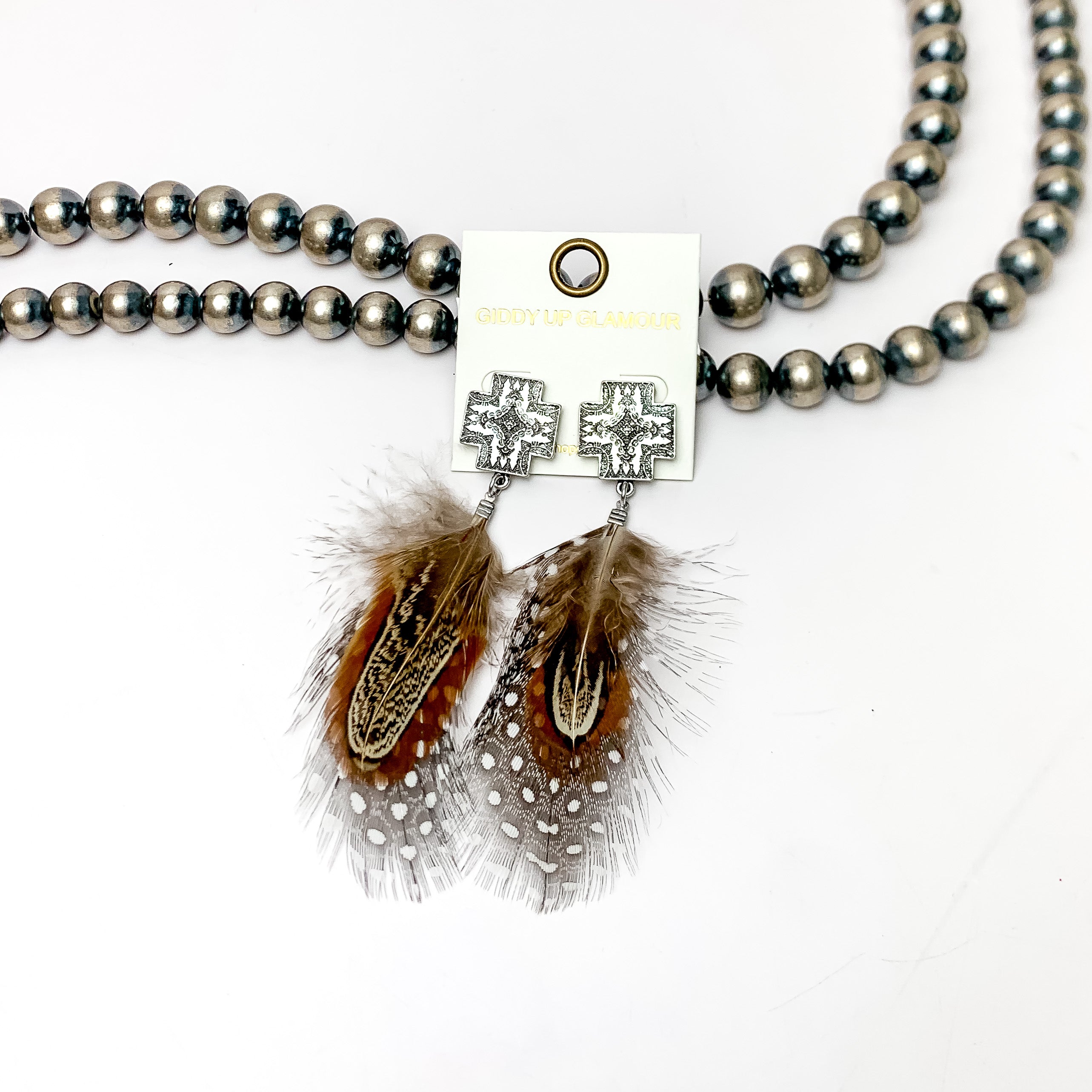 Silver Tone Cross Post Feather Earrings in Brown and Black. These Earrings are pictured laying against Navajo pearls with a white background.