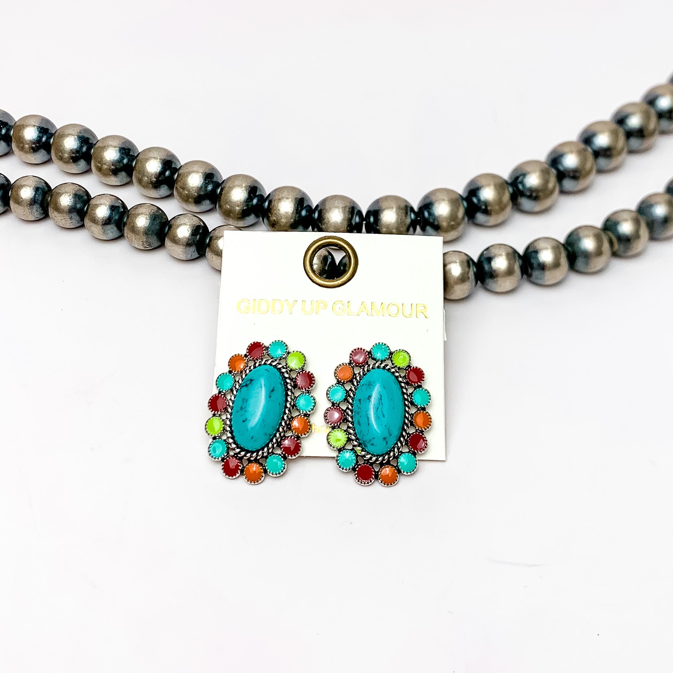 Concho post earrings in multicolor. These earrings are multicolored stones surrounding a bigger turquoise stone. Pictured on a white background with Navajo pearls behind the earrings.
