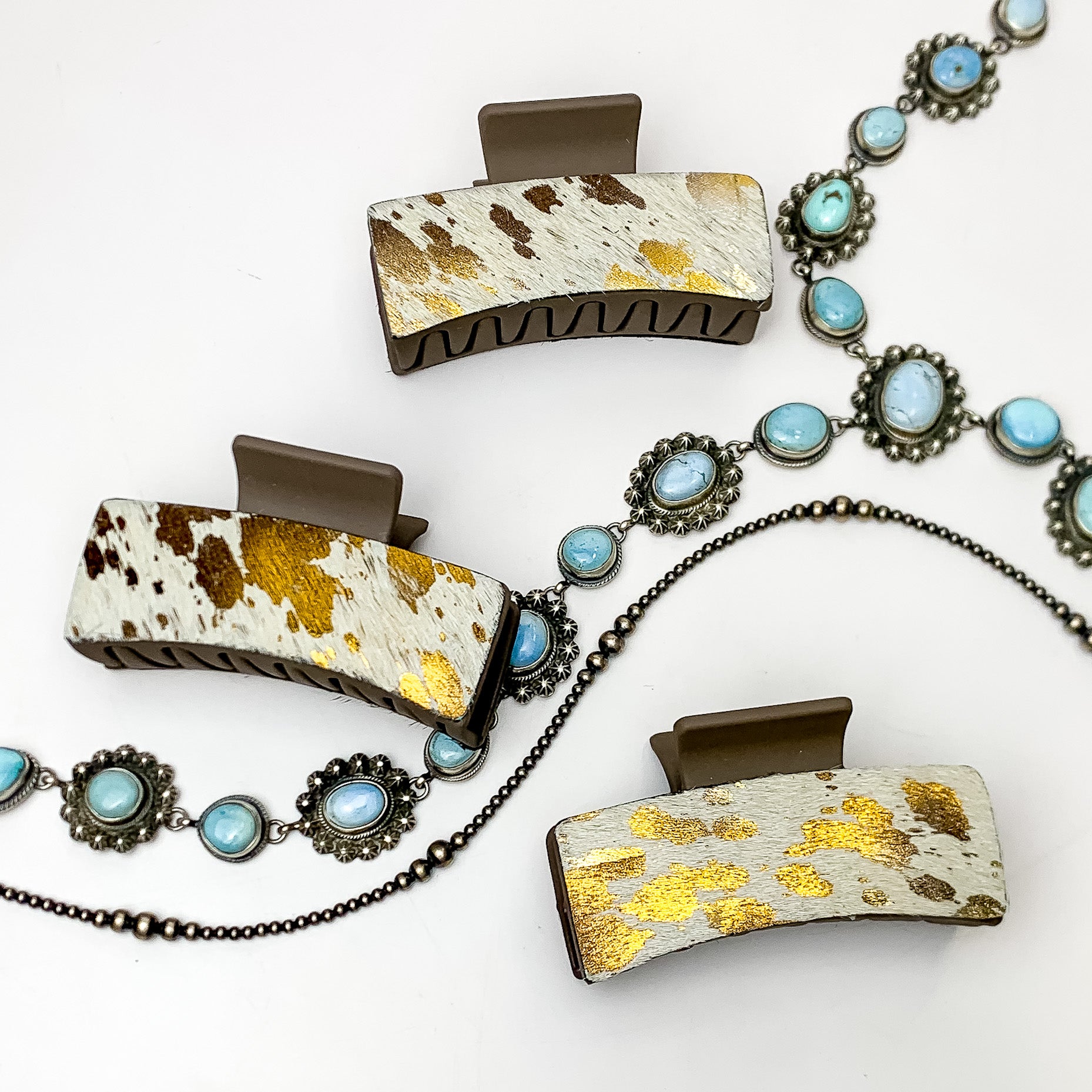 Pictured are three brown hair clips with brown and gold mix hide patches. These earrings are pictured on a white background with silver necklaces under the clips. 