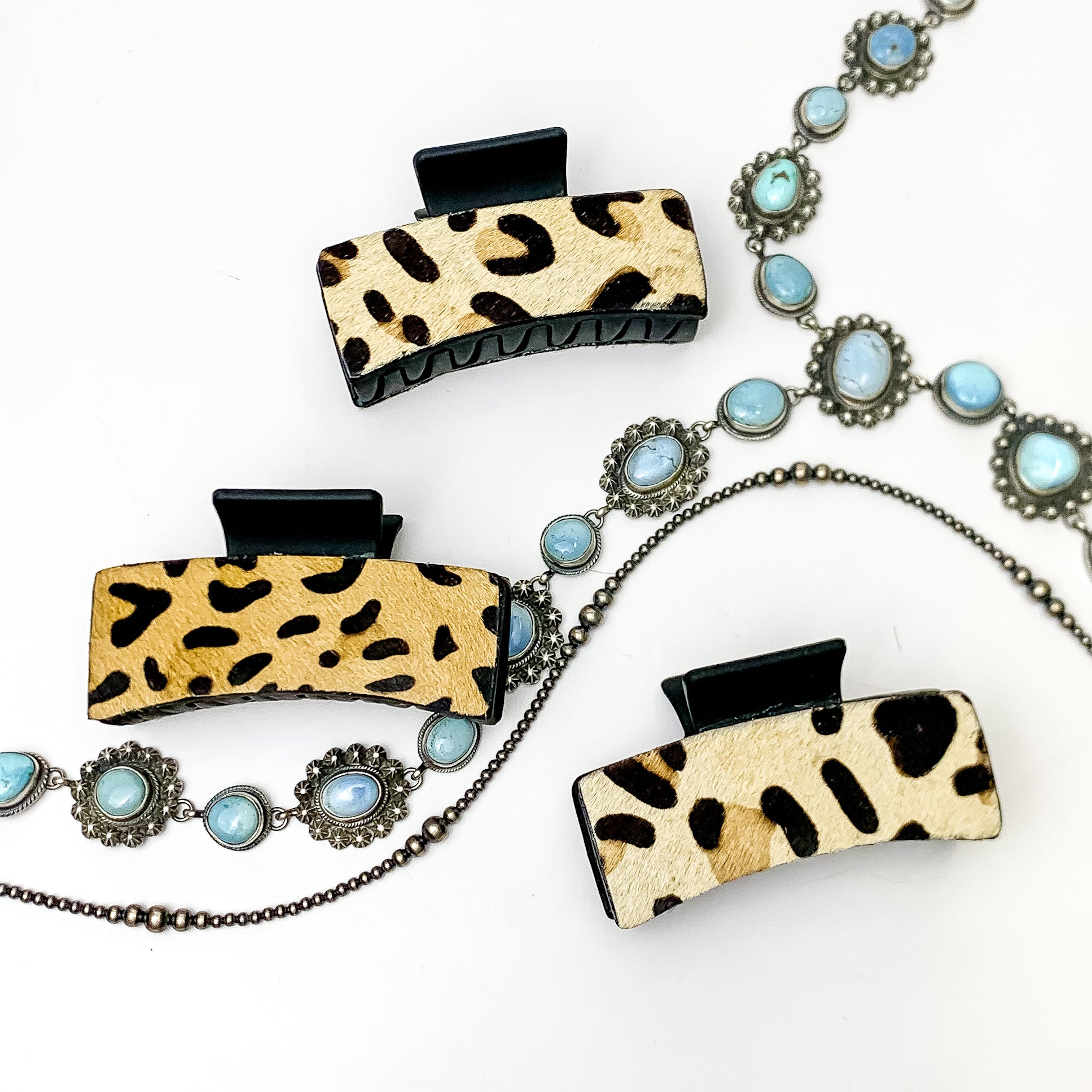 Pictured are three black hair clips with leopard print hide patches. These earrings are pictured on a white background with silver necklaces under the clips. 