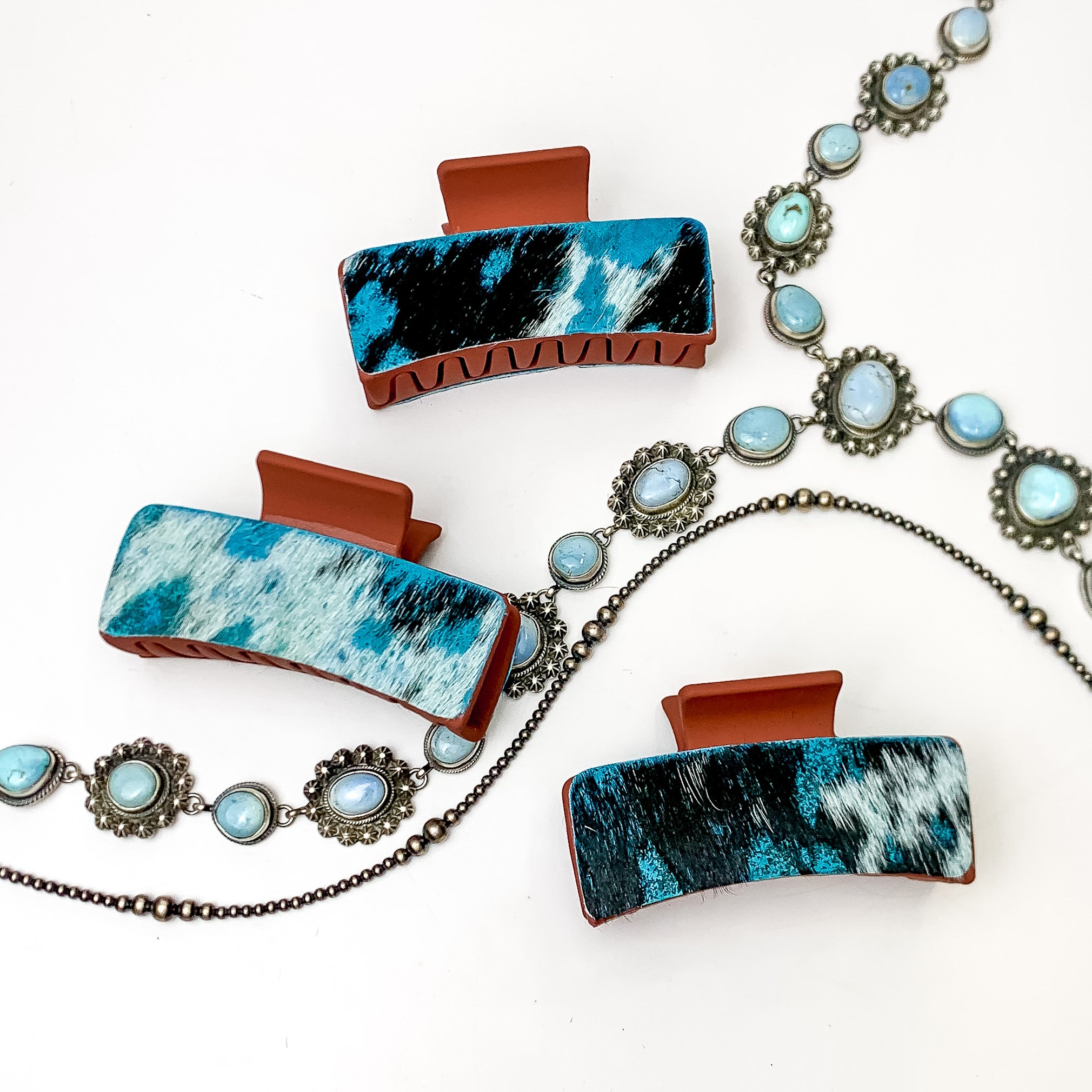 Pictured are three rust red hair clips with turquoise blue mix hide patches. These earrings are pictured on a white background with silver necklaces under the clips. 