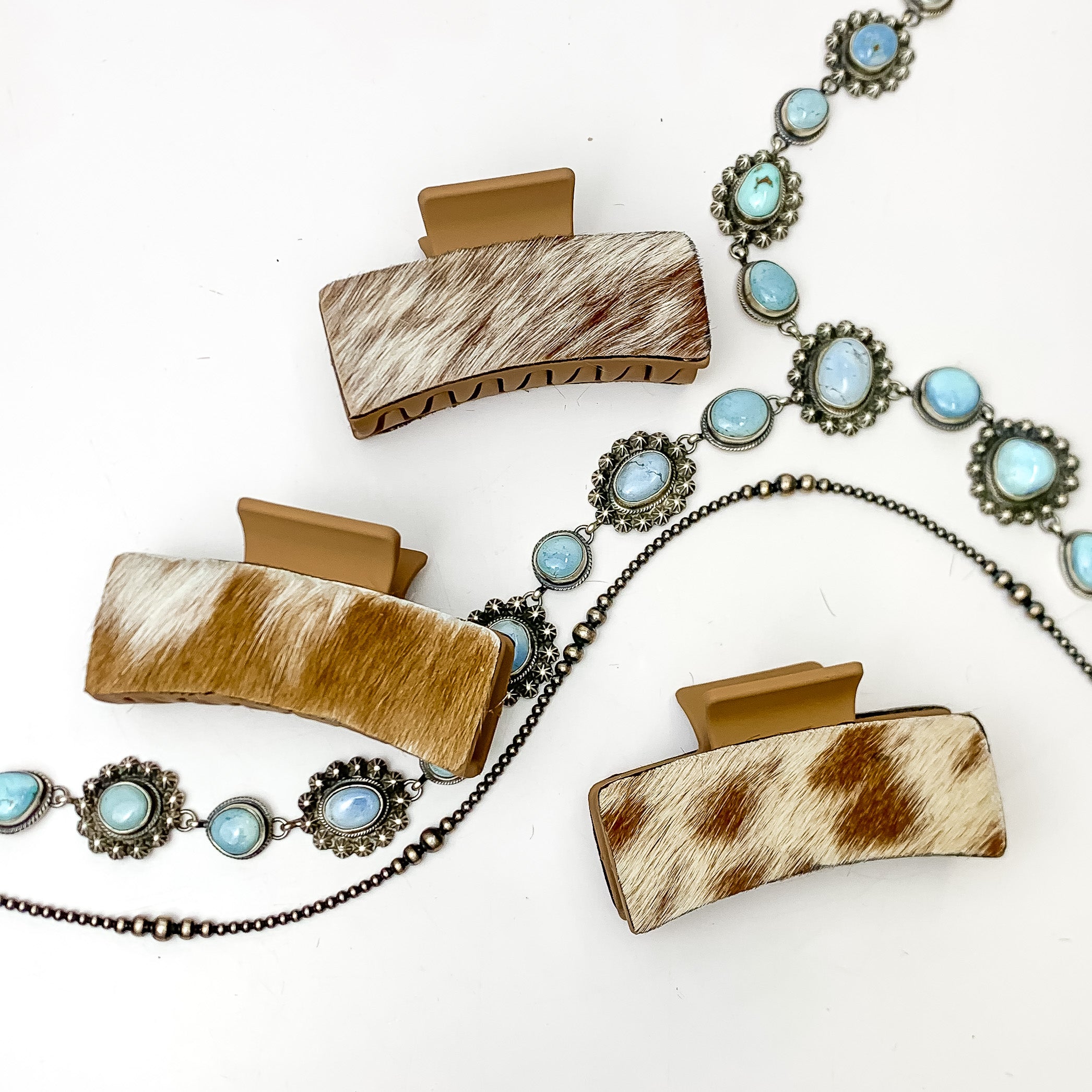 Pictured are three tan hair clips with ivory and tan mix hide patches. These earrings are pictured on a white background with silver necklaces under the clips. 