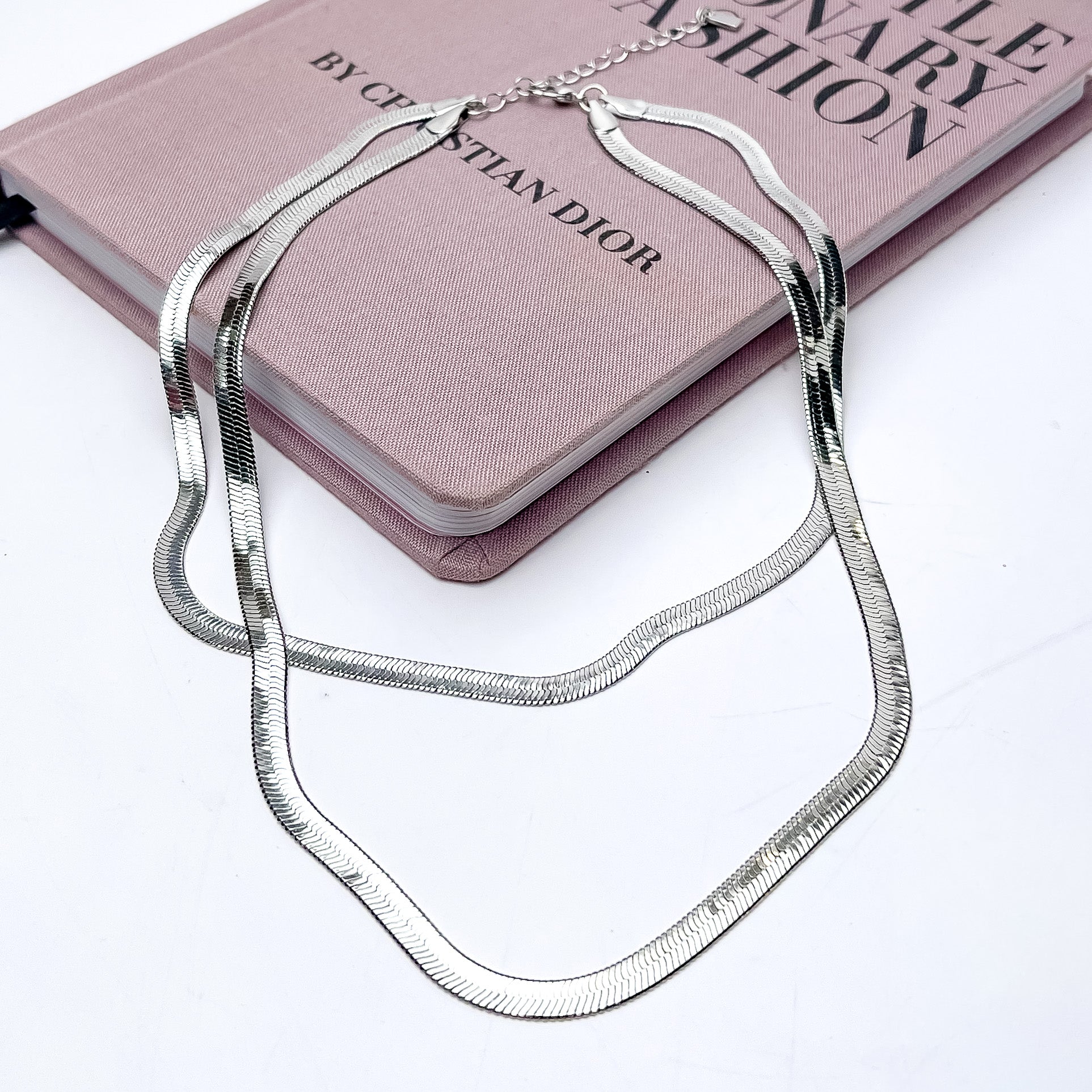 Daily Dose Double Strand Silver Tone Necklace. This necklace is partly laying on a pink book. The background of this picture is white.