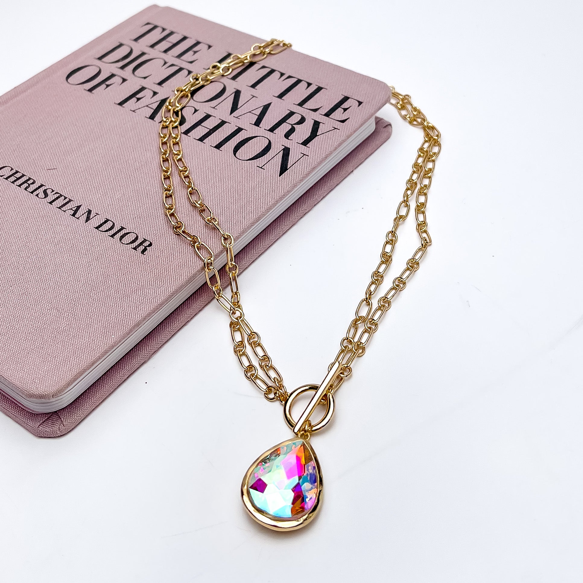 Center of Attention Gold Tone Chain Necklace With AB Crystal Pendant. Pictured on a white background with part of the necklace on a pink book.