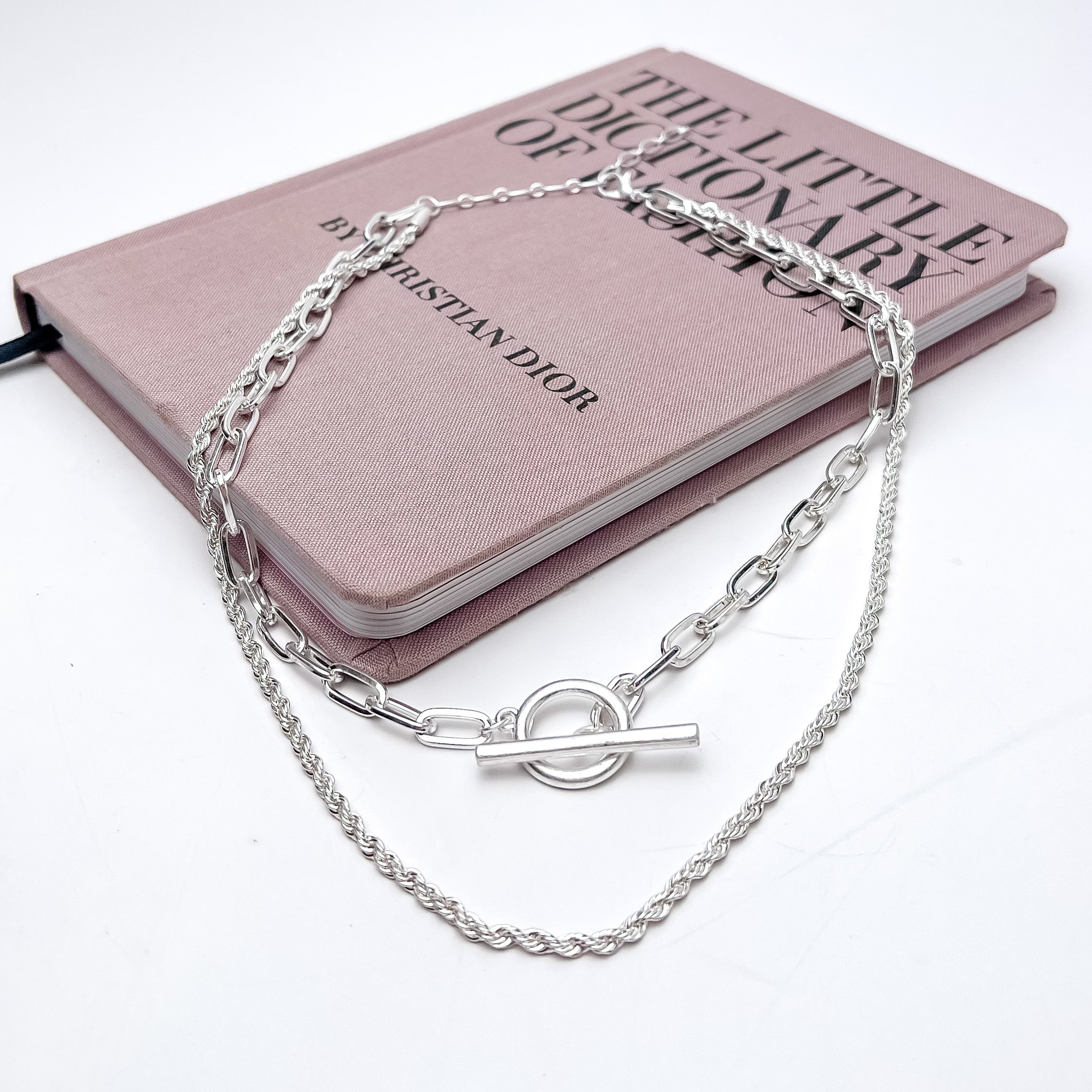 Double Strand Silver Tone Necklace With Front Toggle. This necklace is pictured laying on a pink book with a white background.