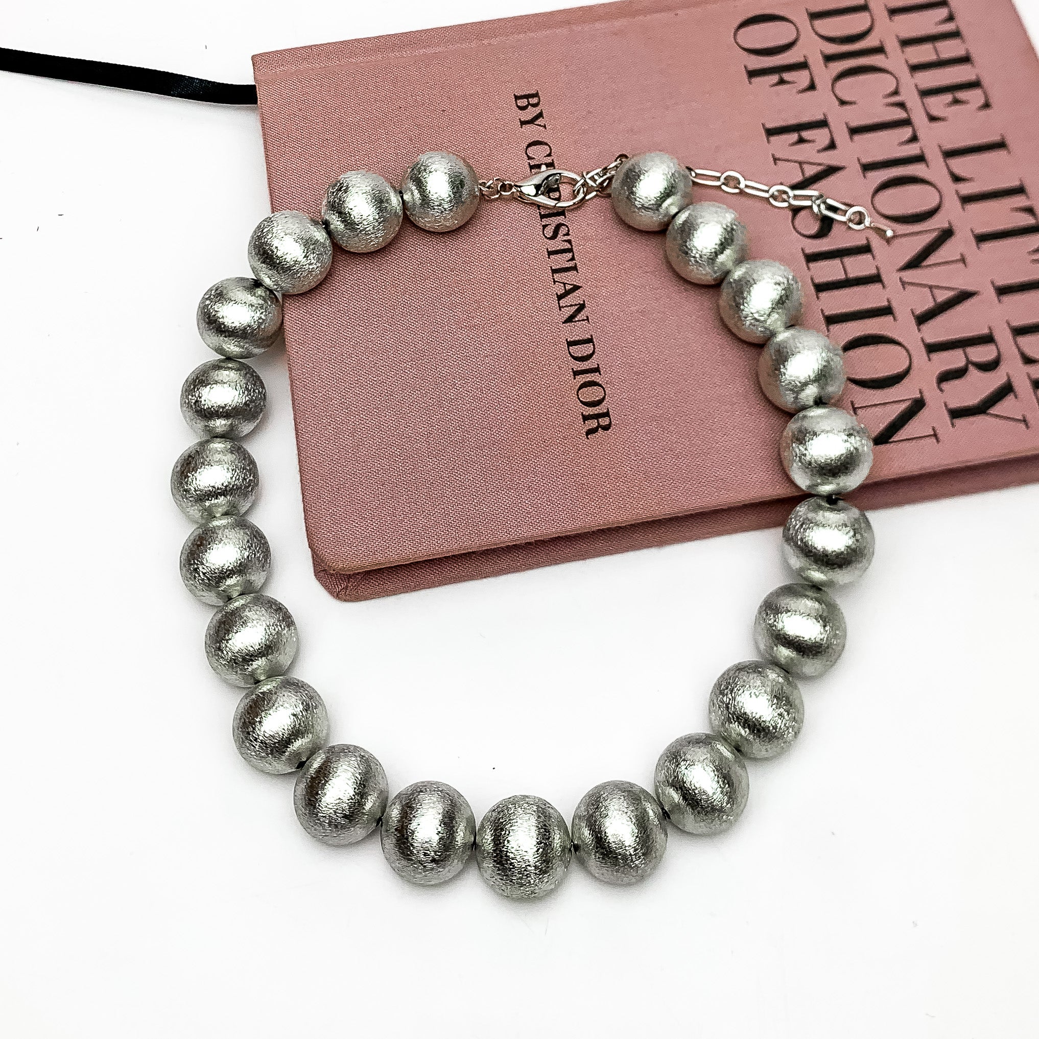 Large Silver Tone Beaded Necklace. This necklace is picktured laying on the side of a pink book with a white background.
