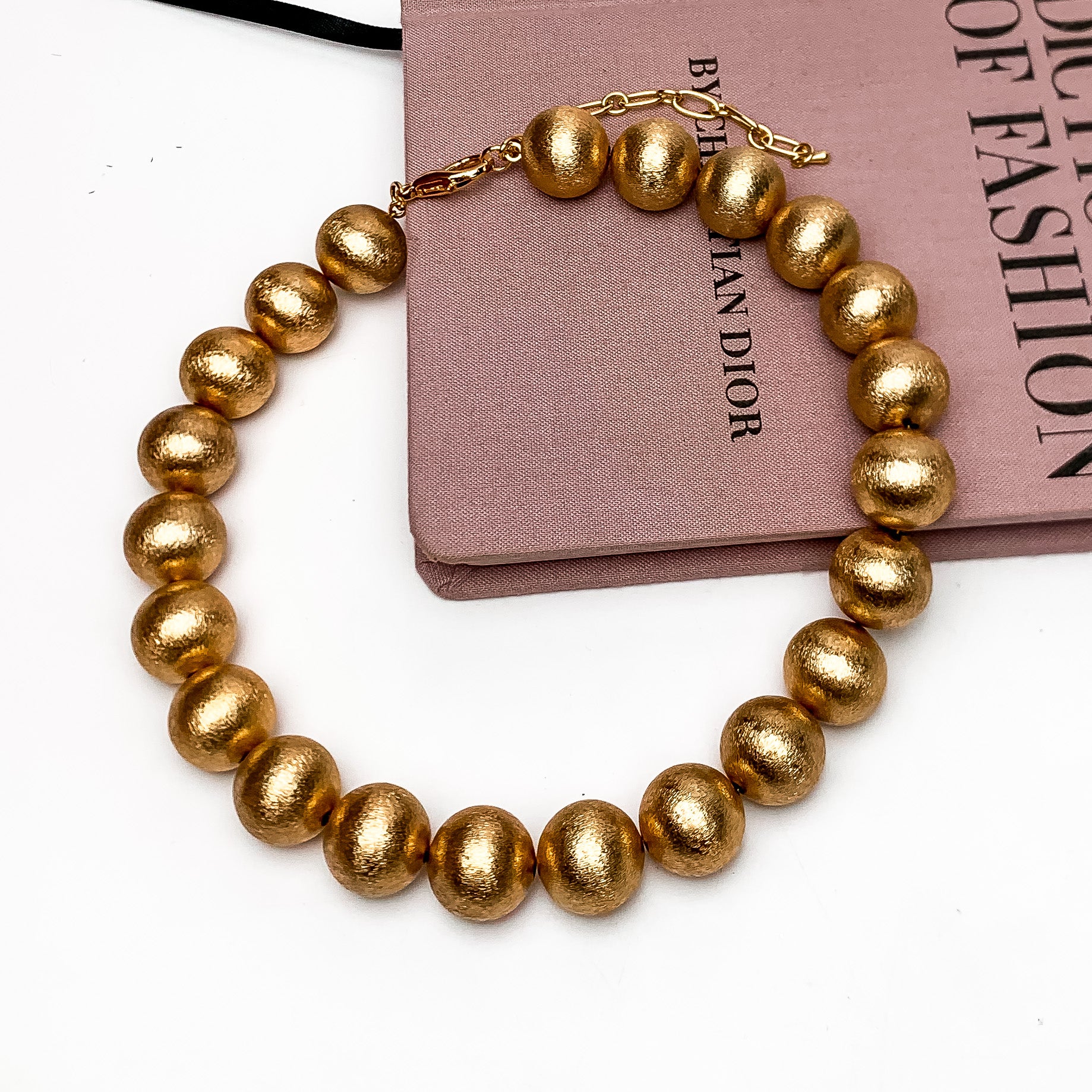 Large Gold Tone Beaded Necklace. This necklace is picktured laying on the side of a pink book with a white background.