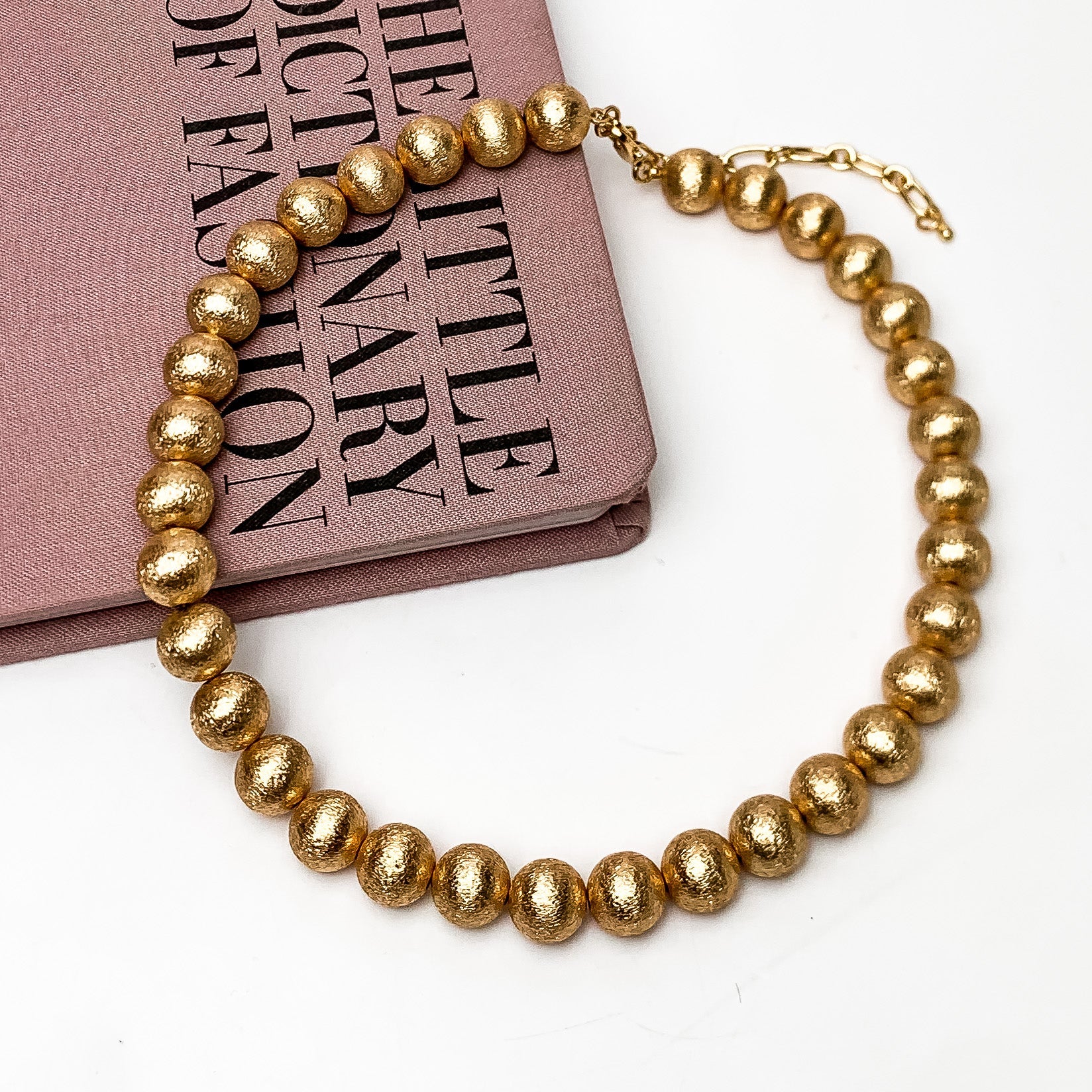 Smaller gold tone beaded necklace. Shiny gold coloring. This necklace is on a pink book with a white background.