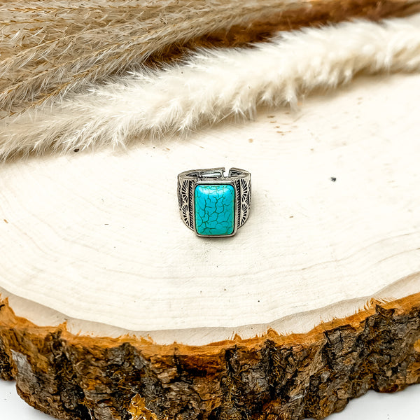 Faux Rectangle Turquoise Stone Stretchy Ring With Silver Tone Design. The ring is pictured on a wood piece with feathers behind it.